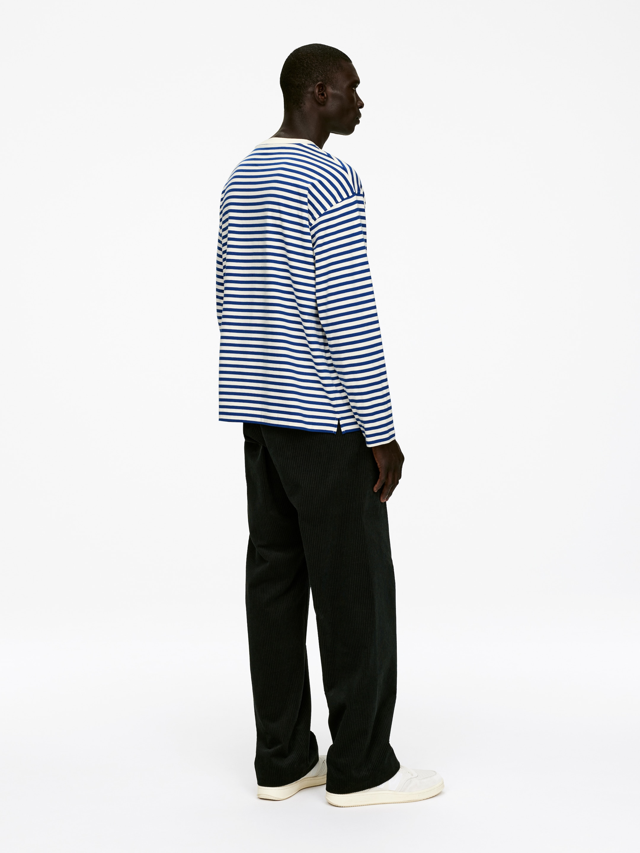Visualizza immagine più grande: Una maglia a maniche lunghe con ampie strisce orizzontali blu e bianche è indossata, caratterizzata da una vestibilità rilassata e un collo a giro. I pantaloni in velluto a coste scuro, a coste larghe, presentano una silhouette rilassata a gamba dritta, e le sneakers basse color bianco sporco completano l'insieme.