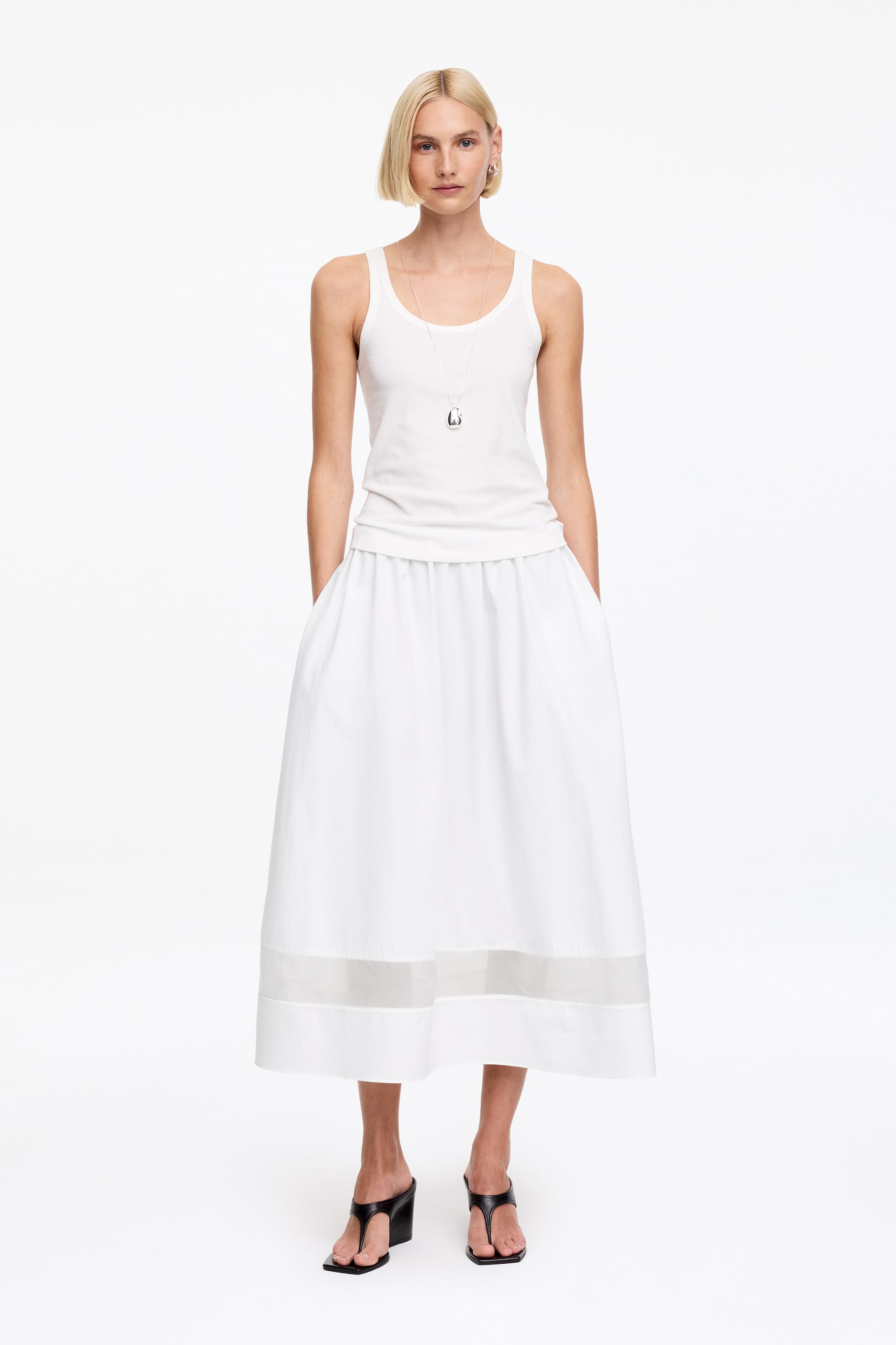 Näytä suurempi kuva: Valkoinen ribbineuletoppi, jossa on pyöreä pääntie ja hihaton muotoilu, on yhdistetty valkoiseen midi-mittaiseen hameeseen. Hameessa on rypytetty vyötärö ja leveä, läpikuultava nauha kiinteän helman yläpuolella. Asua täydentää pitkä hopeinen kaulakoru, jossa on geometrinen riipus, sekä mustat neliökärkiset varvassandaalit korokepohjalla.