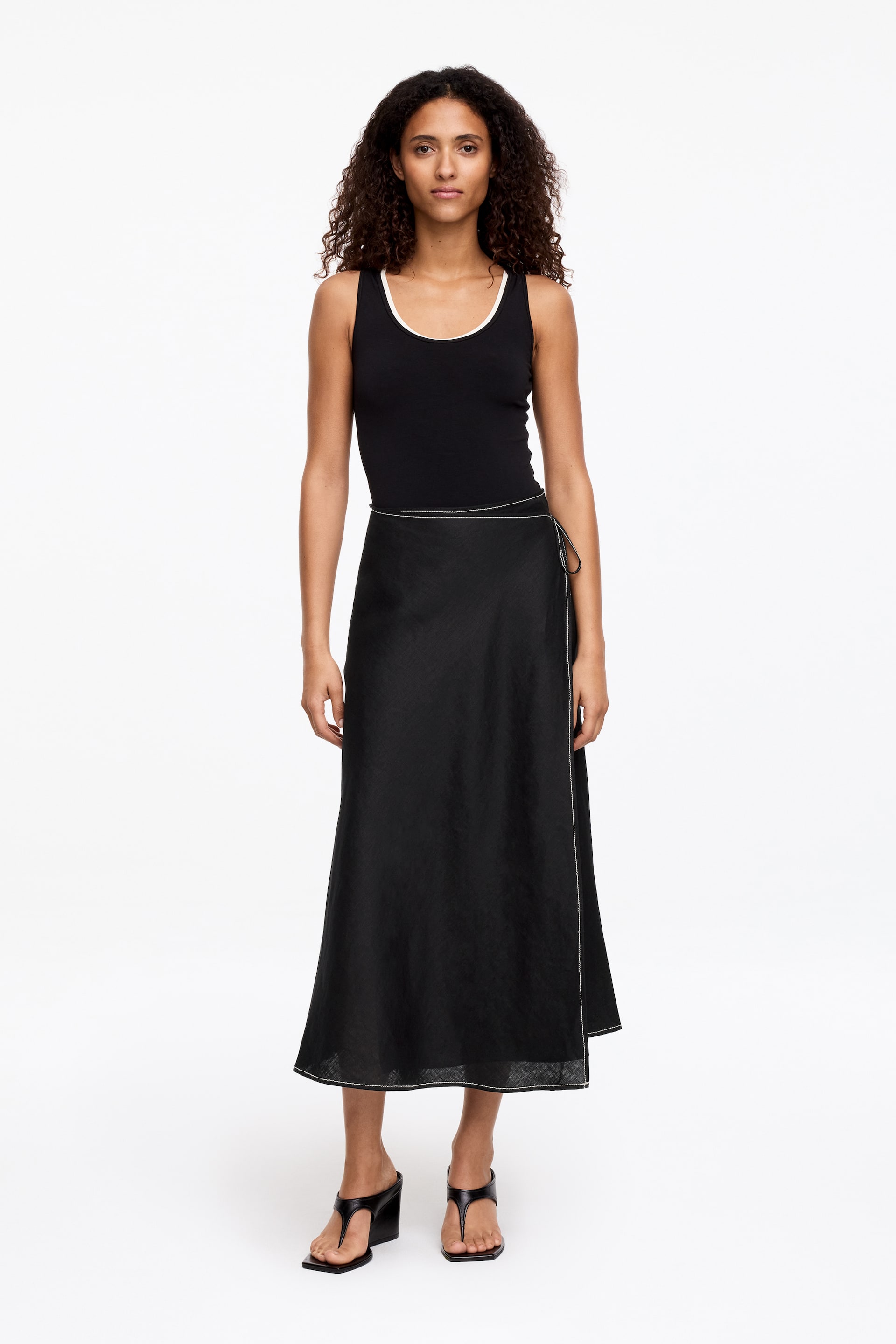 View larger image: A black textured midi wrap skirt featuring white contrast stitching along its wrap opening and hem. It ties at the left hip.