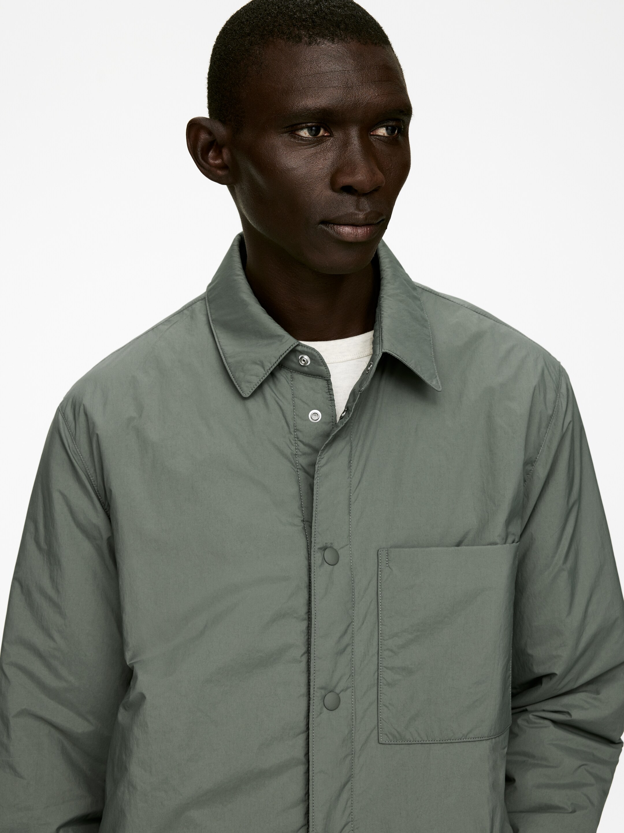Ver imagen más grande: Se está exhibiendo una sobrecamisa acolchada de color verde apagado, con un clásico cuello extendido y un cierre frontal de botones a presión. Muestra un solo bolsillo parche en el pecho izquierdo y está superpuesta sobre un cuello redondo de color claro.