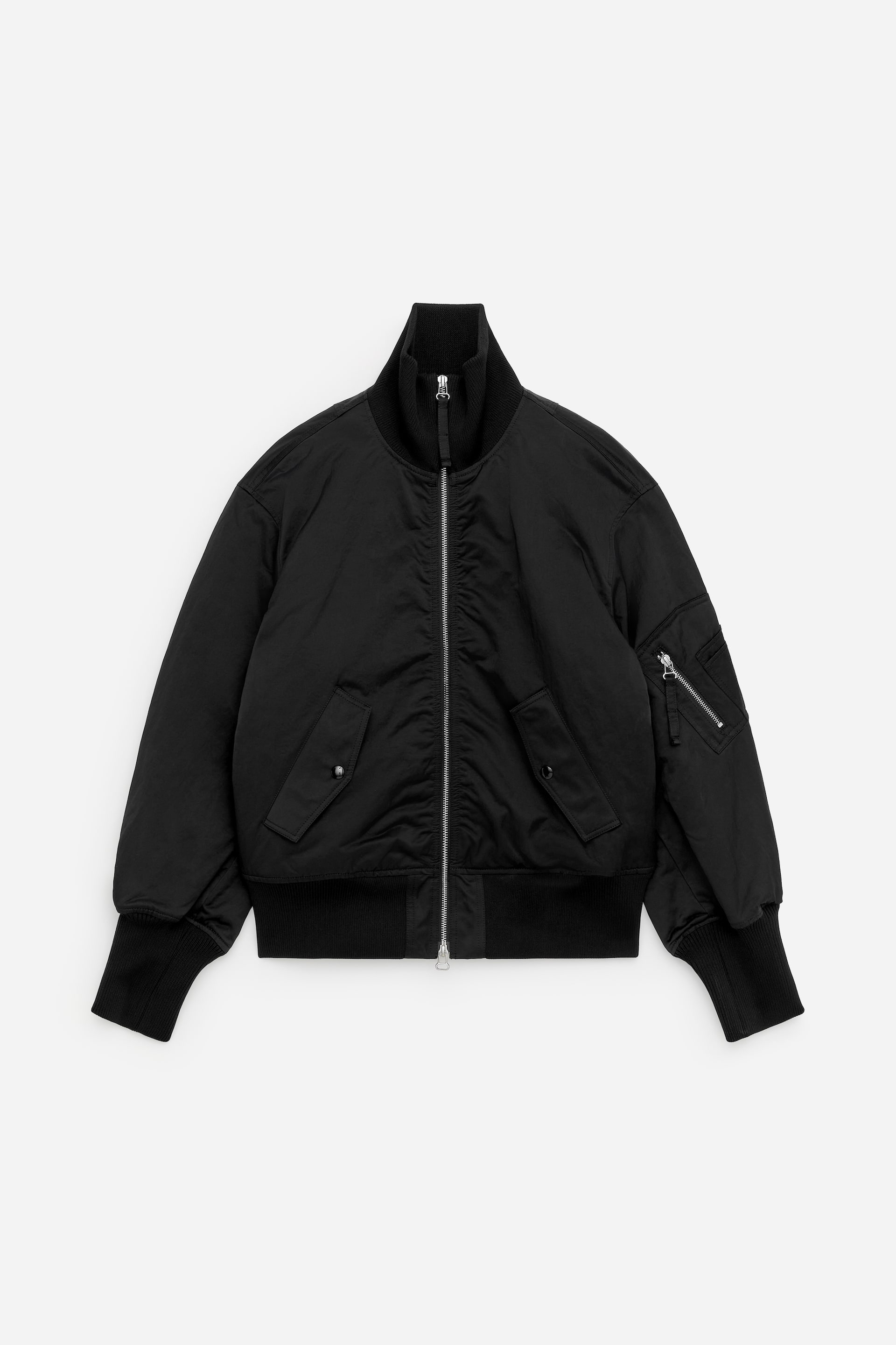 Vis større bilde: FORAN: En svart teknisk nylonjakke med ribbestrikket ståkrage og sentral metallglidelås. Den er designet med ribbedetaljer ved falden og mansjettene, to klafflommer på sidene, og en glidelåslomme på venstre erme.