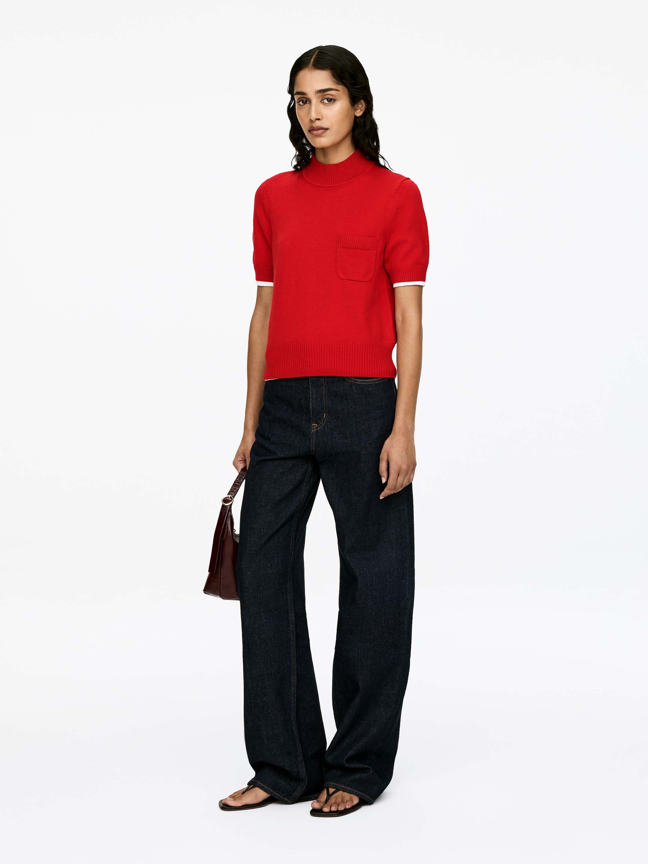 Visa större bild: En röd kortärmad tröja med en mock neck, en ficka på bröstet och ribbade kanter med vita detaljer vid ärmsluten bärs av modellen. Tröjan är matchad med mörka byxor med vida ben.