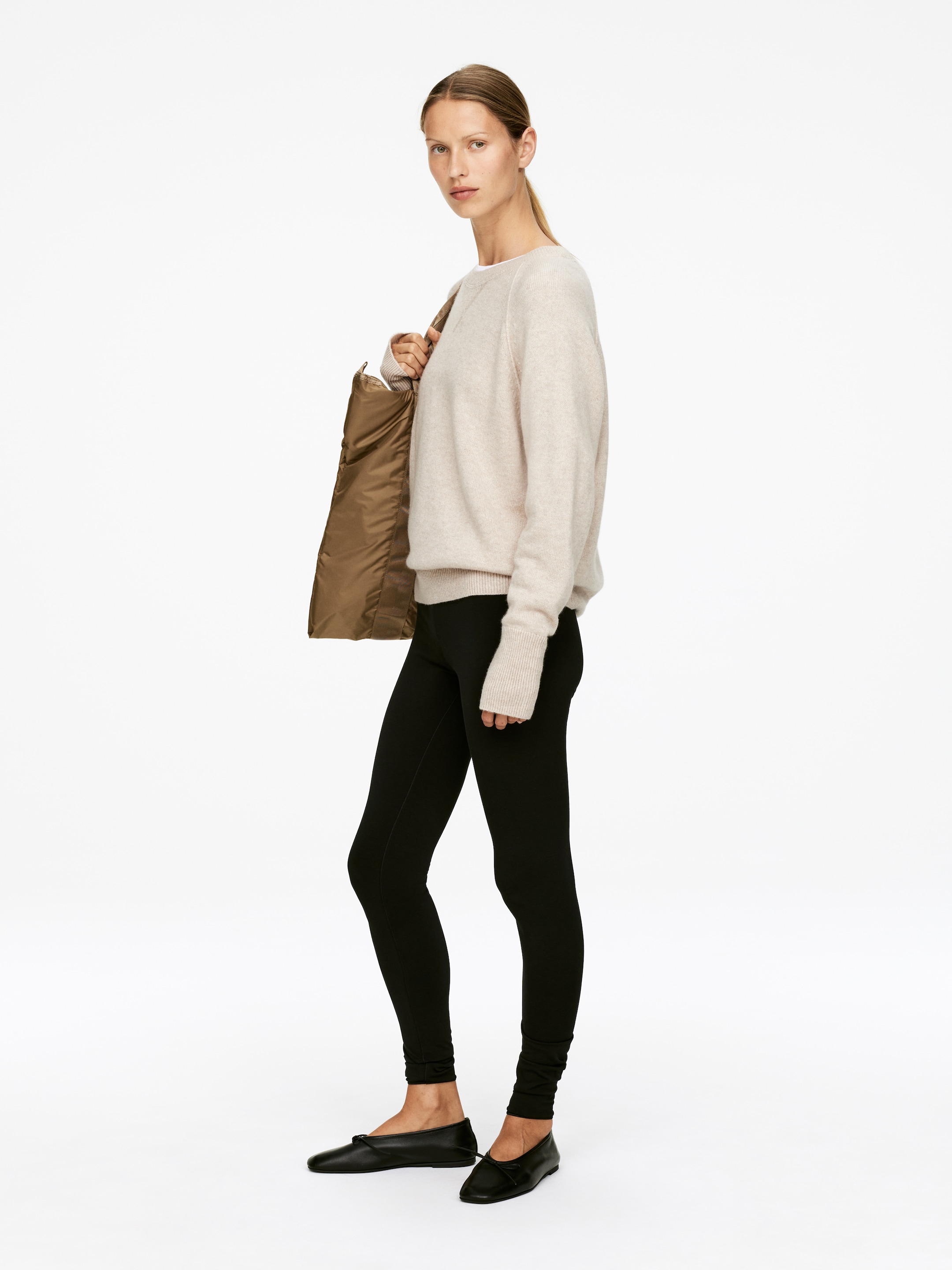 Ver imagen más grande: Un jersey de cuello redondo de color beige claro, de ajuste holgado, con puños y dobladillo acanalados, se presenta desde un ángulo lateral. Se combina con leggings negros de largo completo y zapatos planos negros con un nudo decorativo, mientras que también se lleva un bolso tote marrón suave sobre el hombro.
