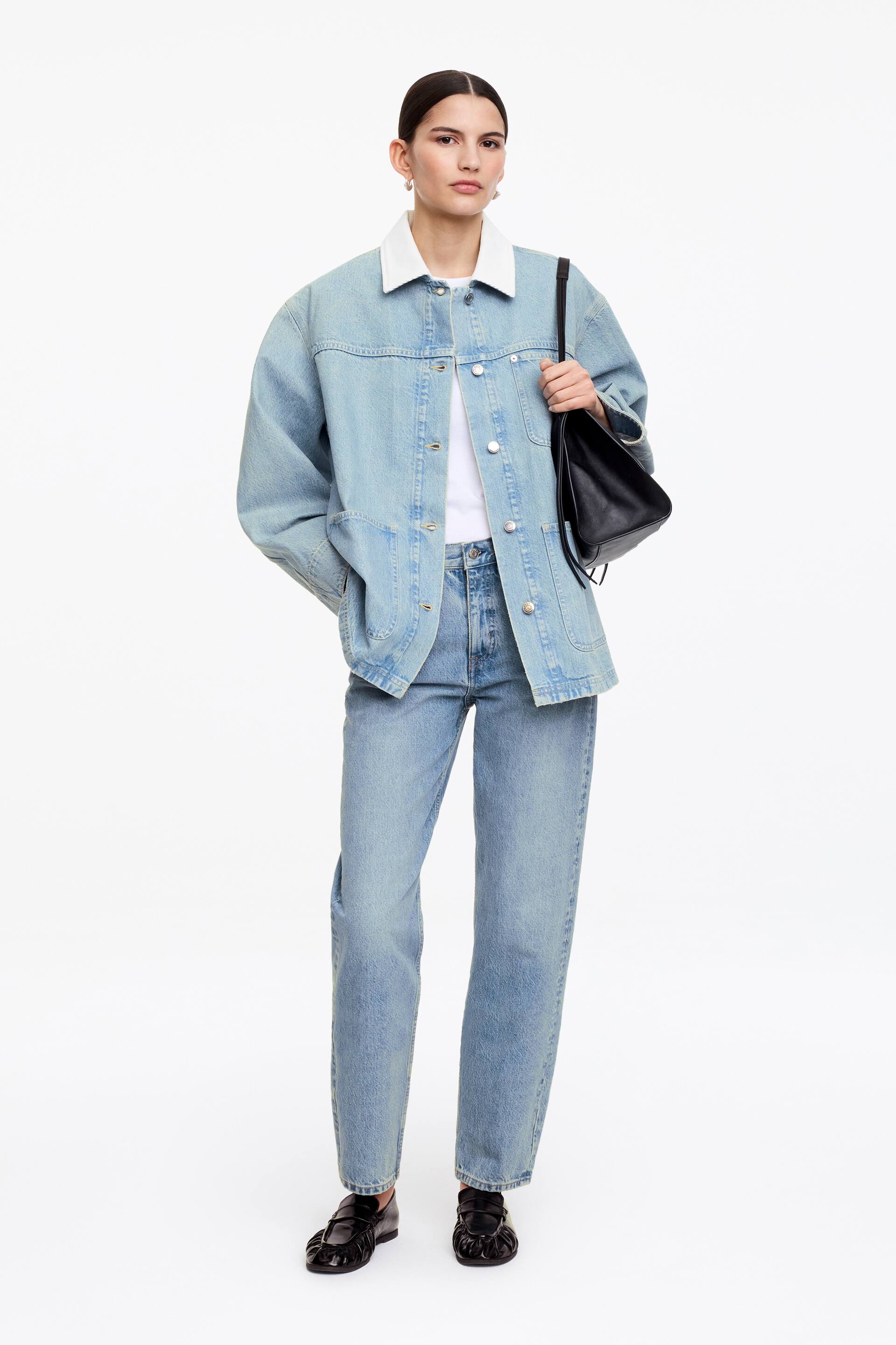 Ver imagen más grande: Una chaqueta vaquera azul claro, con un cuello de pana blanco, cierre frontal con botones de presión y múltiples bolsillos delanteros de parche, está siendo llevada por la modelo. La modelo combina la chaqueta sobre una camiseta blanca, la estiliza con unos vaqueros a juego en azul claro, y lleva un bolso de hombro negro.