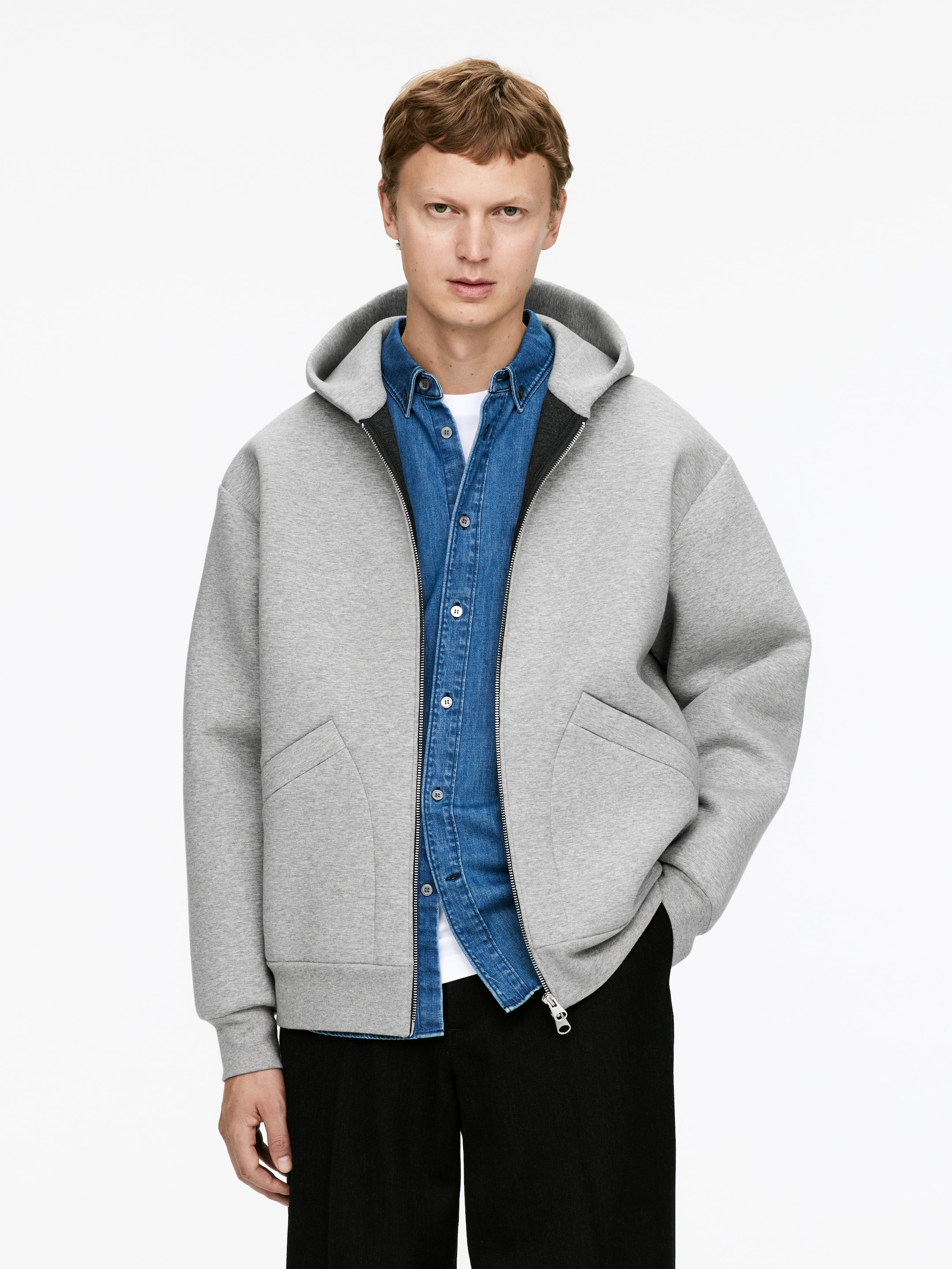 View larger image: A light gray hooded jacket is being shown from the front, featuring a structured, neoprene-like fabric. It is displaying a full-zip front closure with a prominent silver pull, two curved integrated front pockets, and a contrasting dark lining visible at the hood and zipper. The jacket is being worn open over a blue denim shirt.