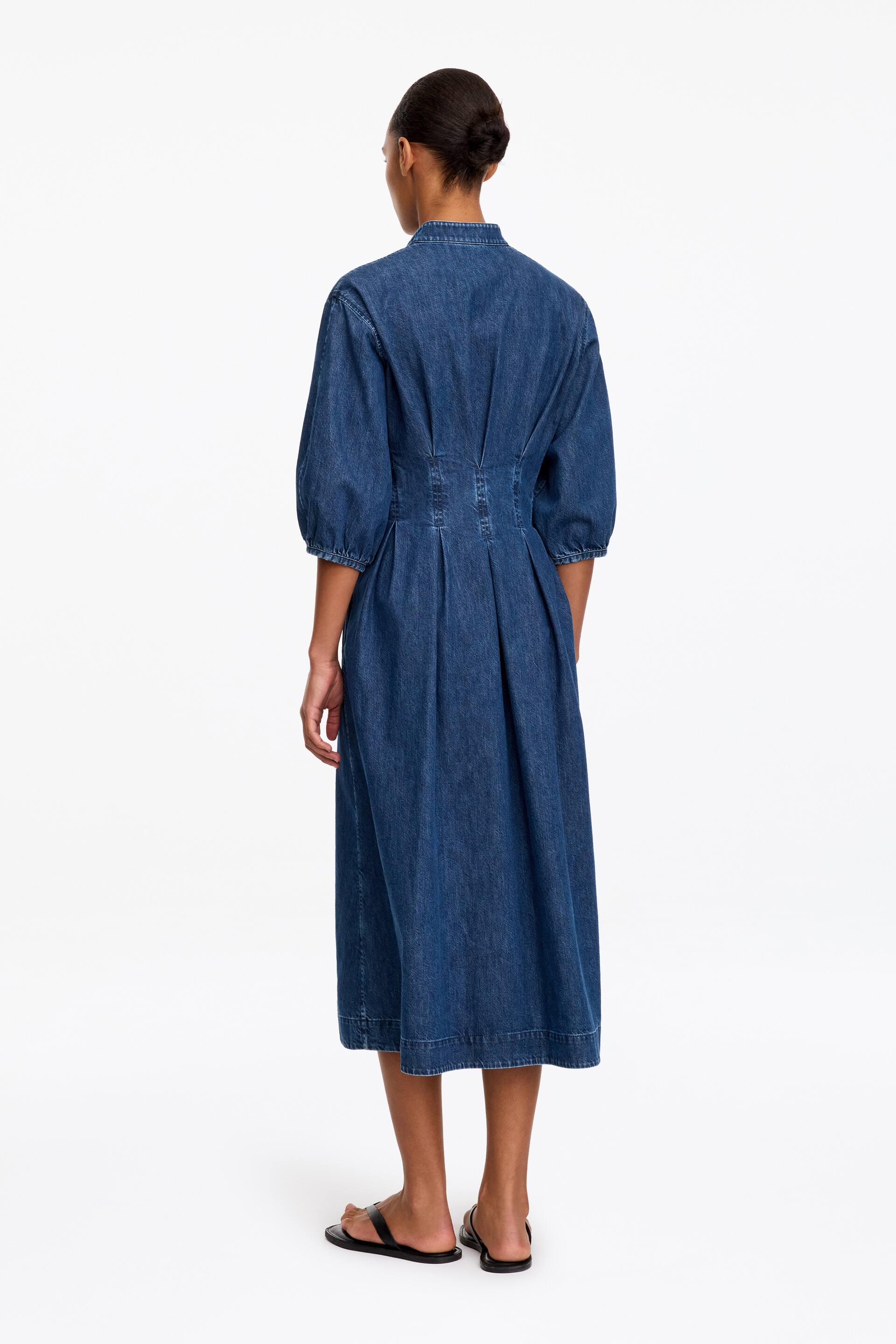 Grotere afbeelding bekijken: Een blauwe denim midi-jurk van katoen wordt gedragen door het model, waarbij de achterkant van het kledingstuk wordt getoond. De jurk heeft een opstaande kraag, gerimpelde mouwen tot aan de elleboog en gestructureerde plooien in de taille die uitlopen in een wijd uitlopende rok.