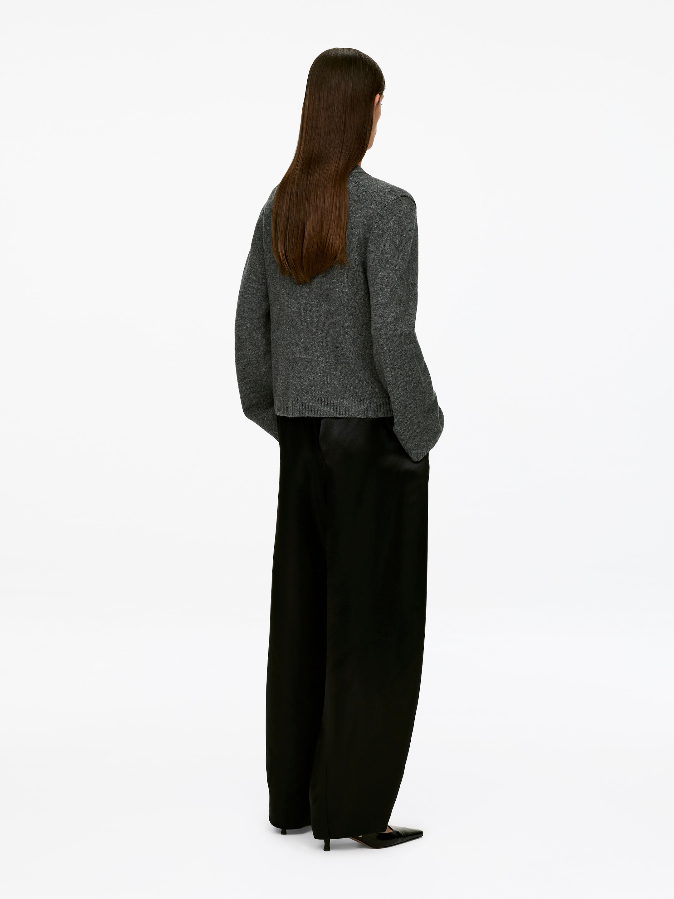 Größeres Bild ansehen: Ein dunkelgrauer Langarm-Cardigan mit geripptem Saum wird vom Model getragen, das mit den Händen in den Taschen schwarzer, weit geschnittener Hosen steht.
