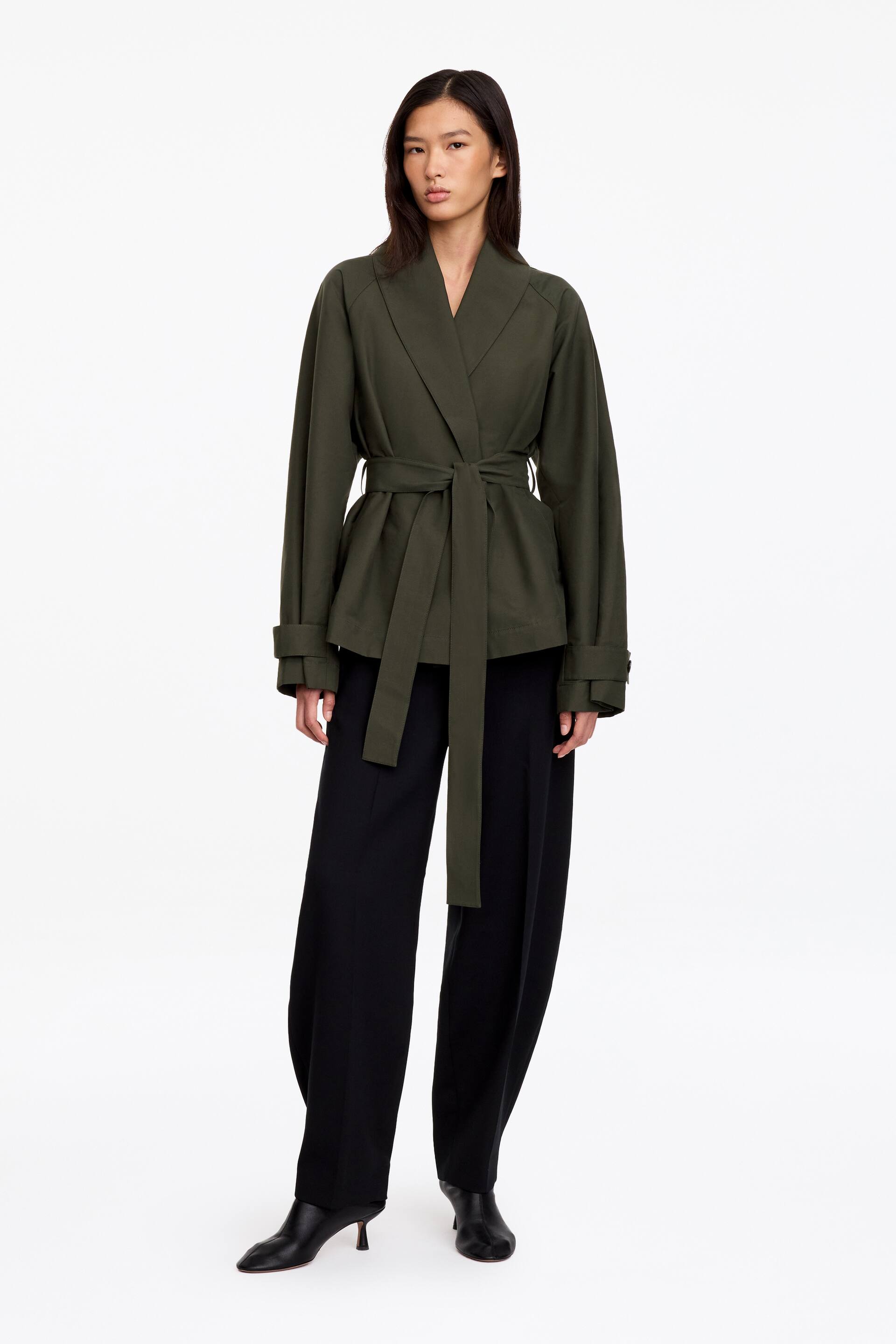 Grösseres Bild ansehen: Eine dunkelolivgrüne Jacke mit Schalkragen, Raglanärmeln und Ärmellaschen wird vom Model getragen und ist in der Taille mit einem passenden Bindegürtel aus dem gleichen Stoff geschnürt. Die Jacke wird mit schwarzen Wide-Leg-Hosen und schwarzen Stiefeletten mit spitzer Spitze kombiniert.
