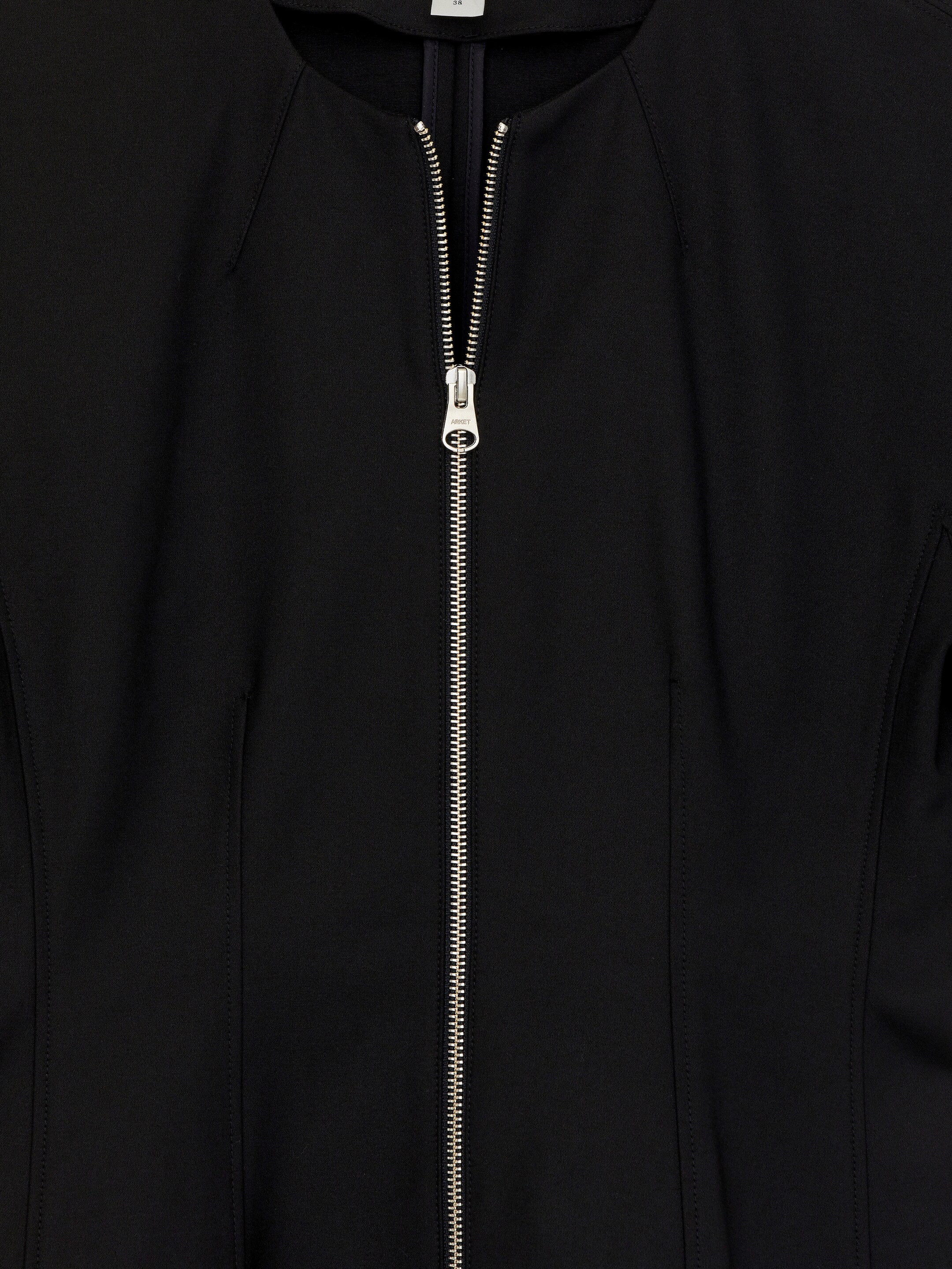 Visa större bild: En svart peplum-topp presenteras framifrån, med en rund halsringning, raglanärmar och en tvåvägs silverfärgad dragkedja framtill som är delvis uppdragen från toppen och avslöjar ett mörkt foder. Vertikala sömmar formar den smala passformen på livstycket och den åtsittande midjan.