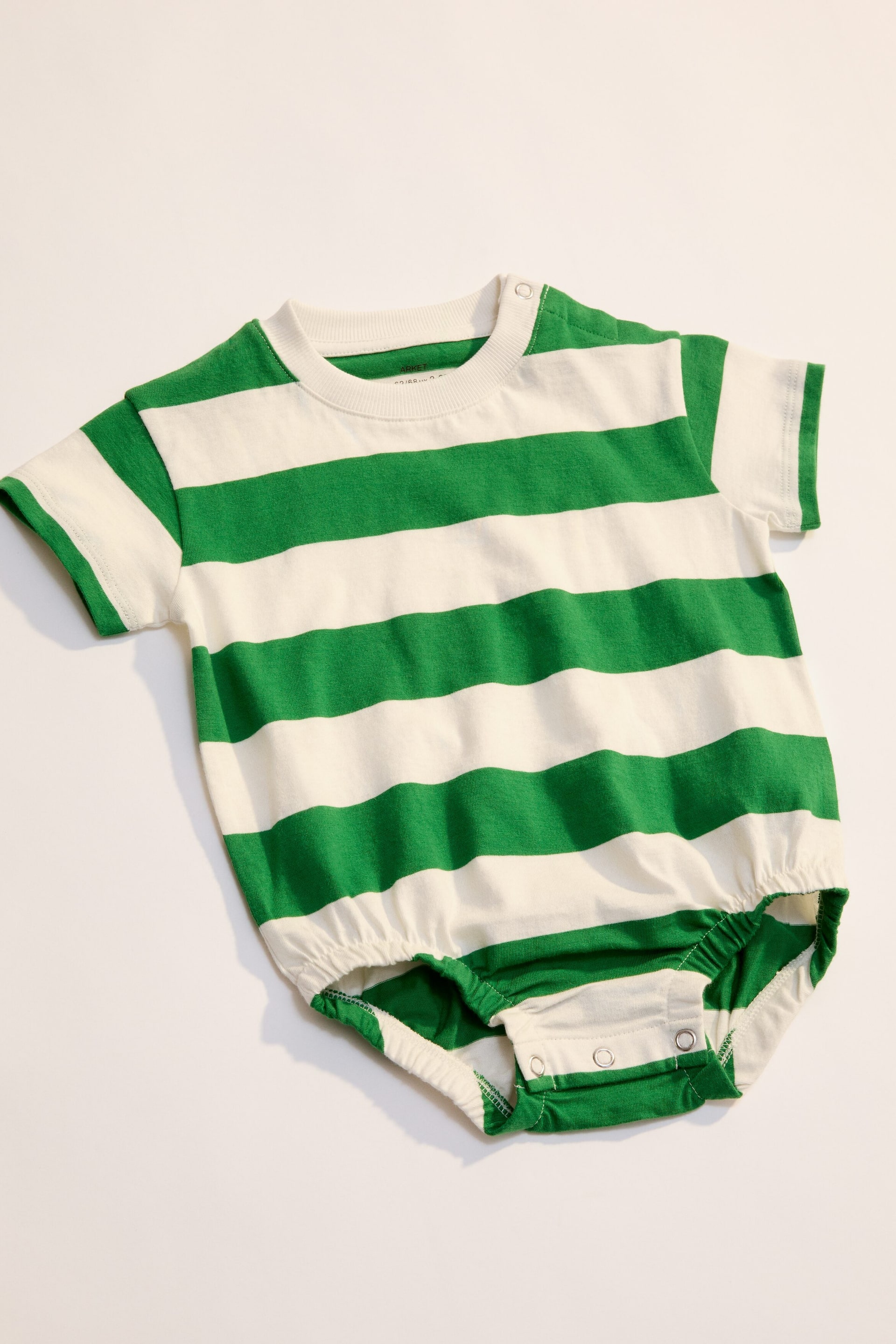 Visa större bild: En babyromper ligger platt och visar sin framsida med gröna och off-white horisontella ränder. Den har korta ärmar och en ribbad rund halsringning, med en tryckknappsstängning synlig på vänster axel. Resår samlar benöppningarna, och tryckknappar fäster grenen.