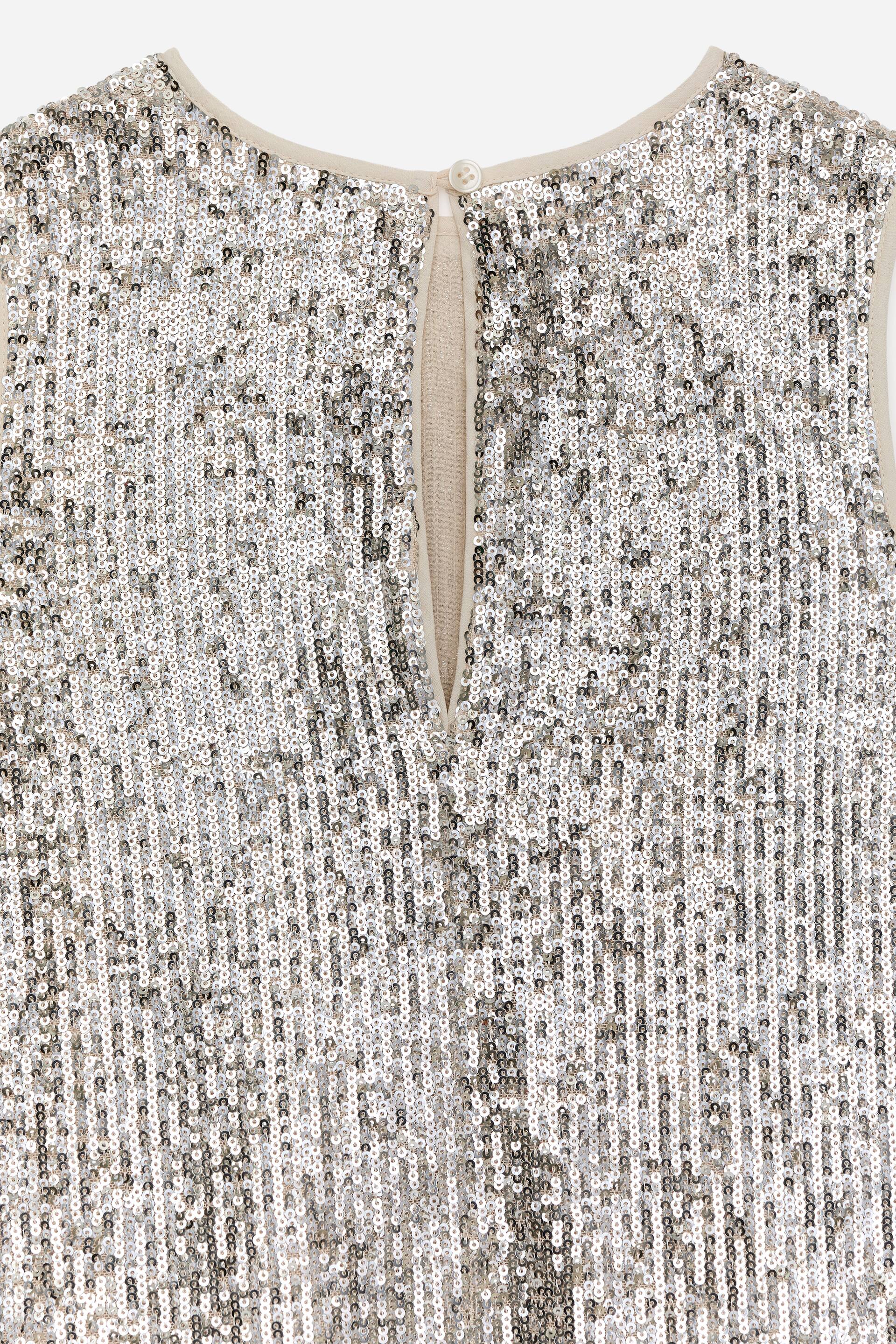 Agrandir l'image: Un vêtement sans manches en sequins argentés présente un décolleté rond et une ouverture en trou de serrure dans le haut du dos. L'ouverture se ferme avec un seul bouton blanc cassé et une boucle, et une bordure beige clair termine le décolleté, les emmanchures et l'ouverture en trou de serrure.
