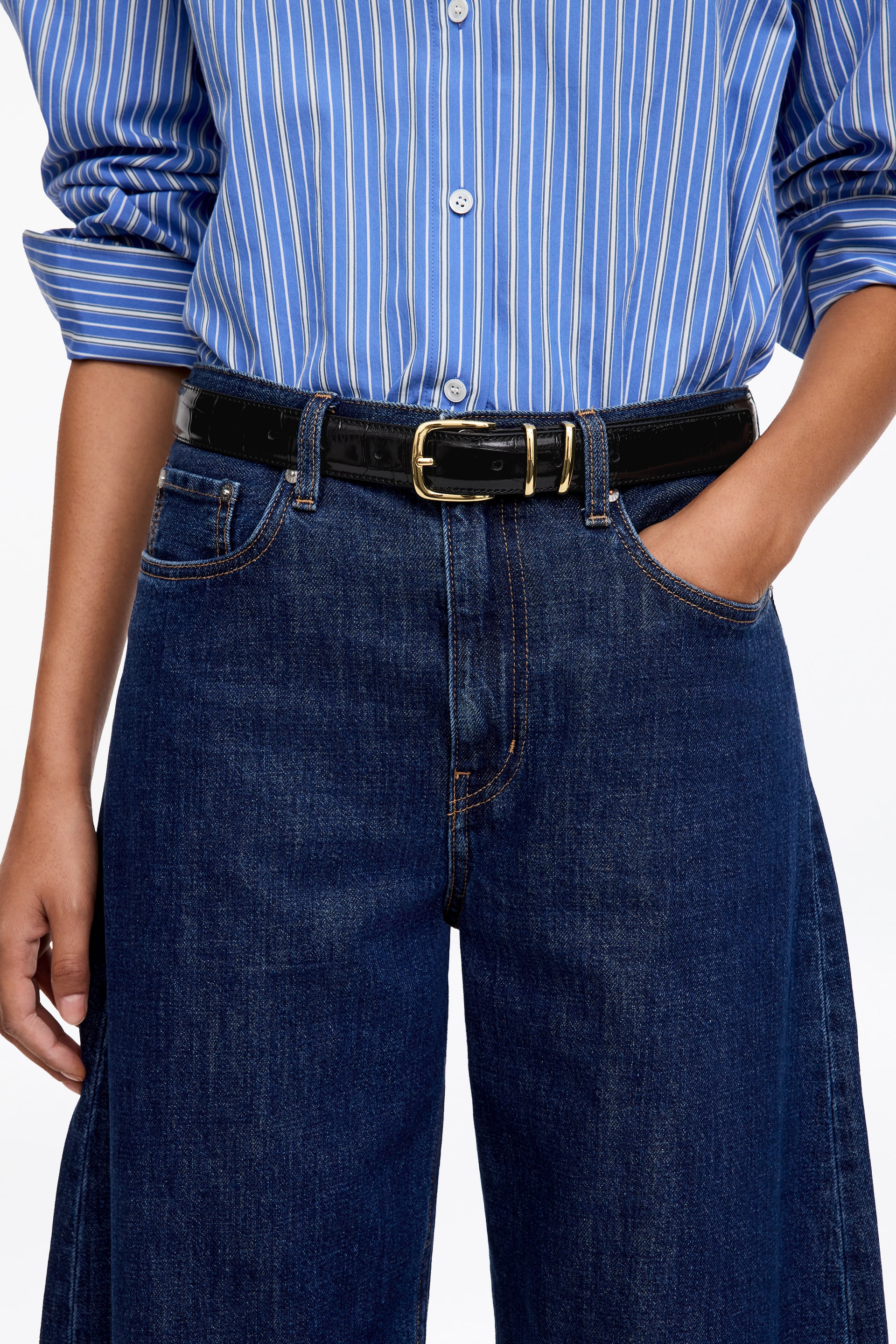 Ver imagen más grande: Un cinturón de cuero negro texturizado, de 30 mm de ancho, con una hebilla de tono dorado, se lleva alrededor de la cintura de la modelo. Sujeta unos vaqueros de denim azul oscuro y una camisa a rayas azules y blancas.