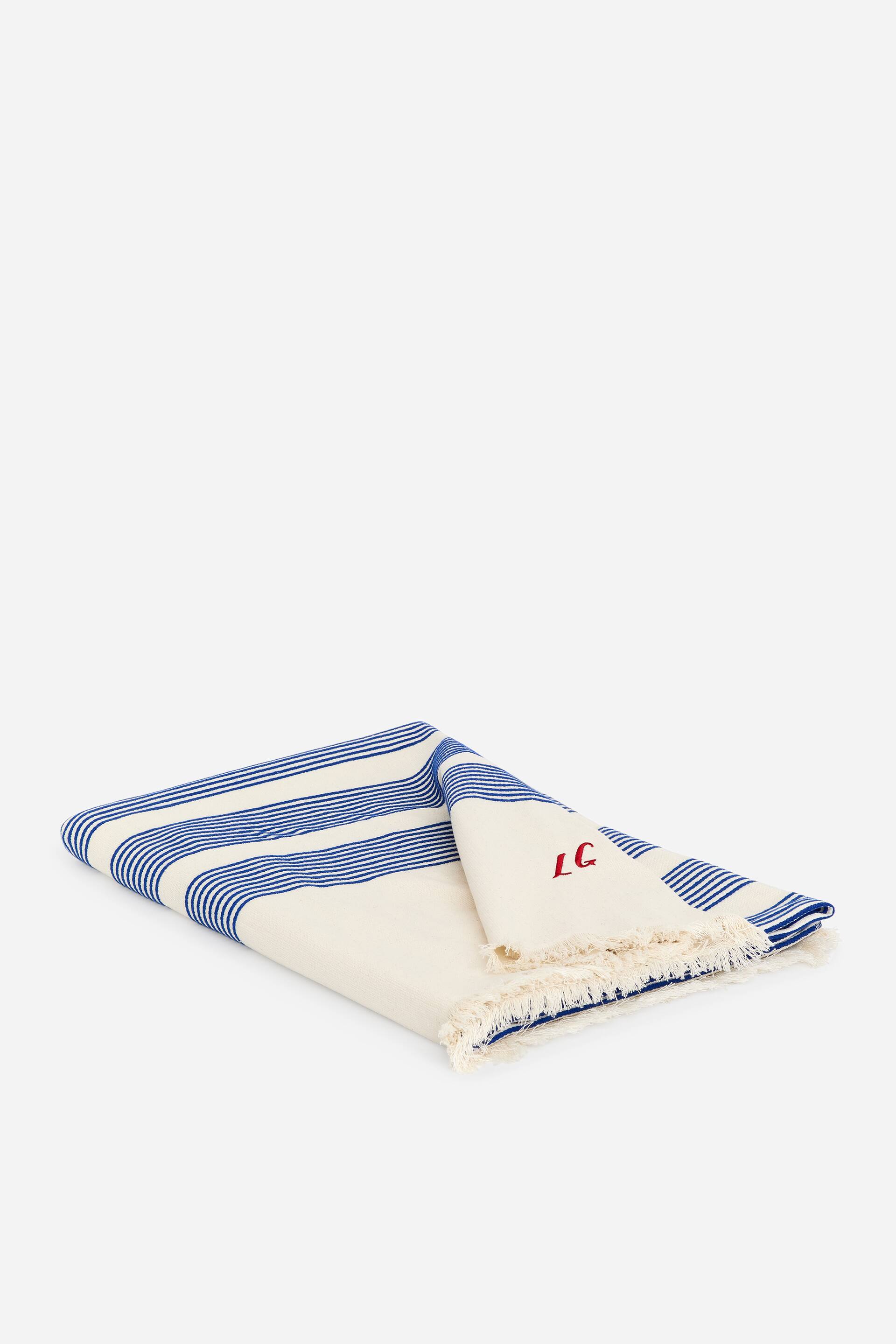 Grotere afbeelding bekijken: De ARKET en Laila Gohar Monogram Deken verschijnt gevouwen, met een crèmekleurige stof met horizontale blauwe strepen. Het heeft een rood geborduurd 'LC' monogram in een hoek en rafelige randen.