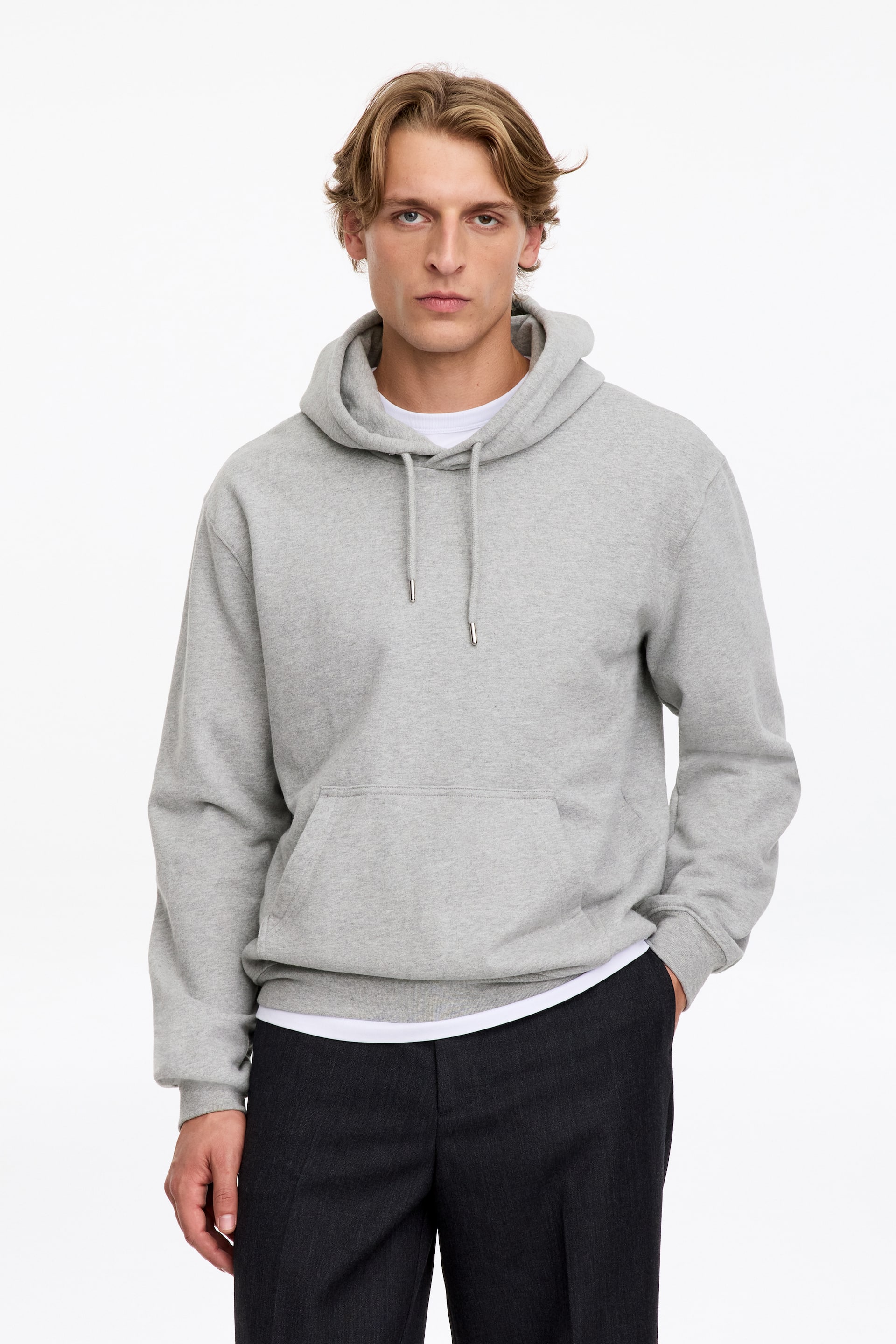 Agrandir l'image: Un sweat à capuche gris clair, doté de cordons de serrage, d’une poche frontale et de finitions côtelées, est porté par le mannequin. Ce sweat à la coupe régulière est superposé à un t-shirt blanc et associé à un pantalon foncé.