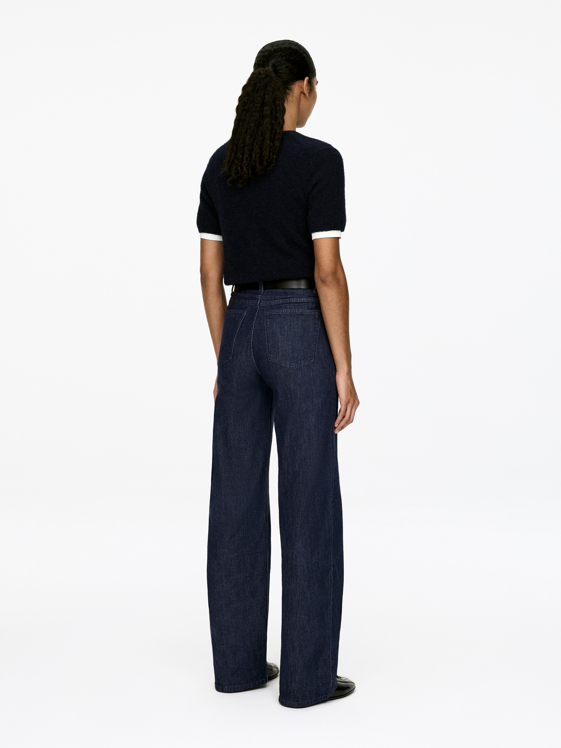 Agrandir l'image: Un haut tricoté à manches courtes bleu foncé, avec des poignets blancs, est porté rentré dans un jean à jambes larges de couleur foncée, vu de dos. Le jean présente deux poches arrière visibles et une fine ceinture sombre serre la taille.