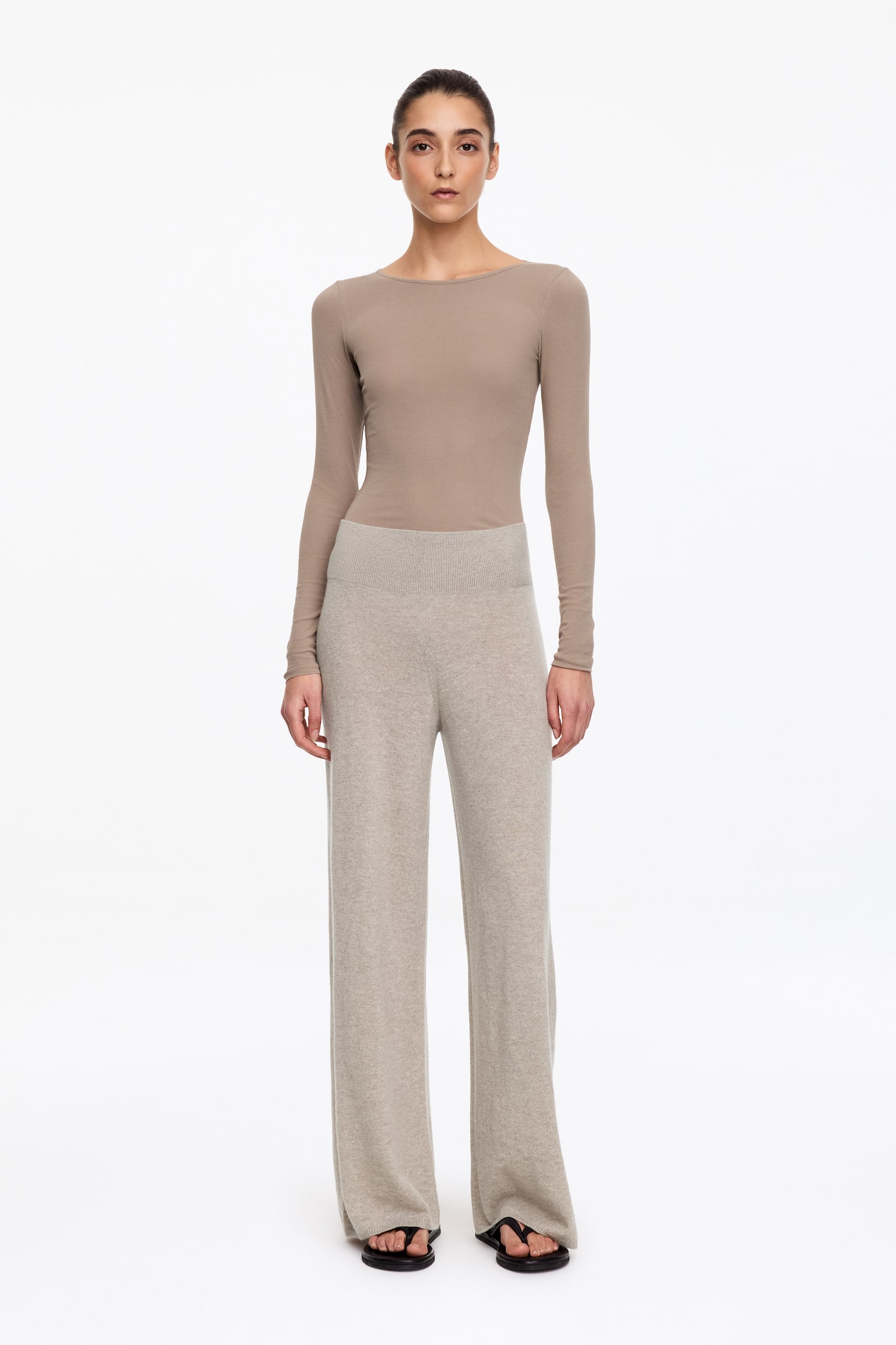 Een lichtbeige gebreide broek staat van voren afgebeeld en heeft een wijde pijp en een hoge, geribde tailleband. De broek toont een fijne breistructuur met rechte, netjes afgewerkte zomen. - 1