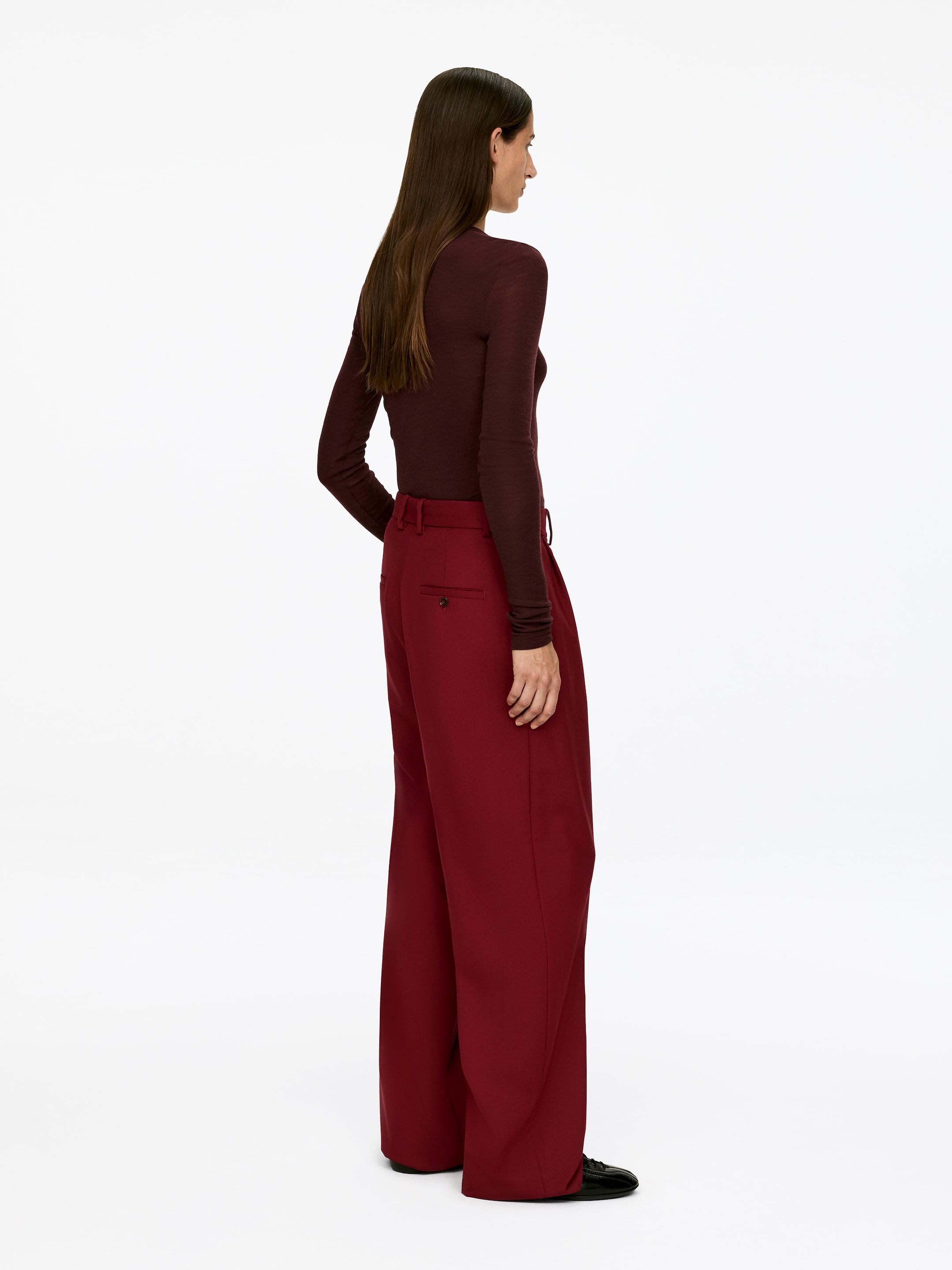 Grösseres Bild ansehen: Die tiefroten Hopfensack-Wollhosen, die eine hohe Taille und weite Beine aufweisen, werden vom Modell getragen, dessen rechte Hand in der Nähe des Bunds ruht.