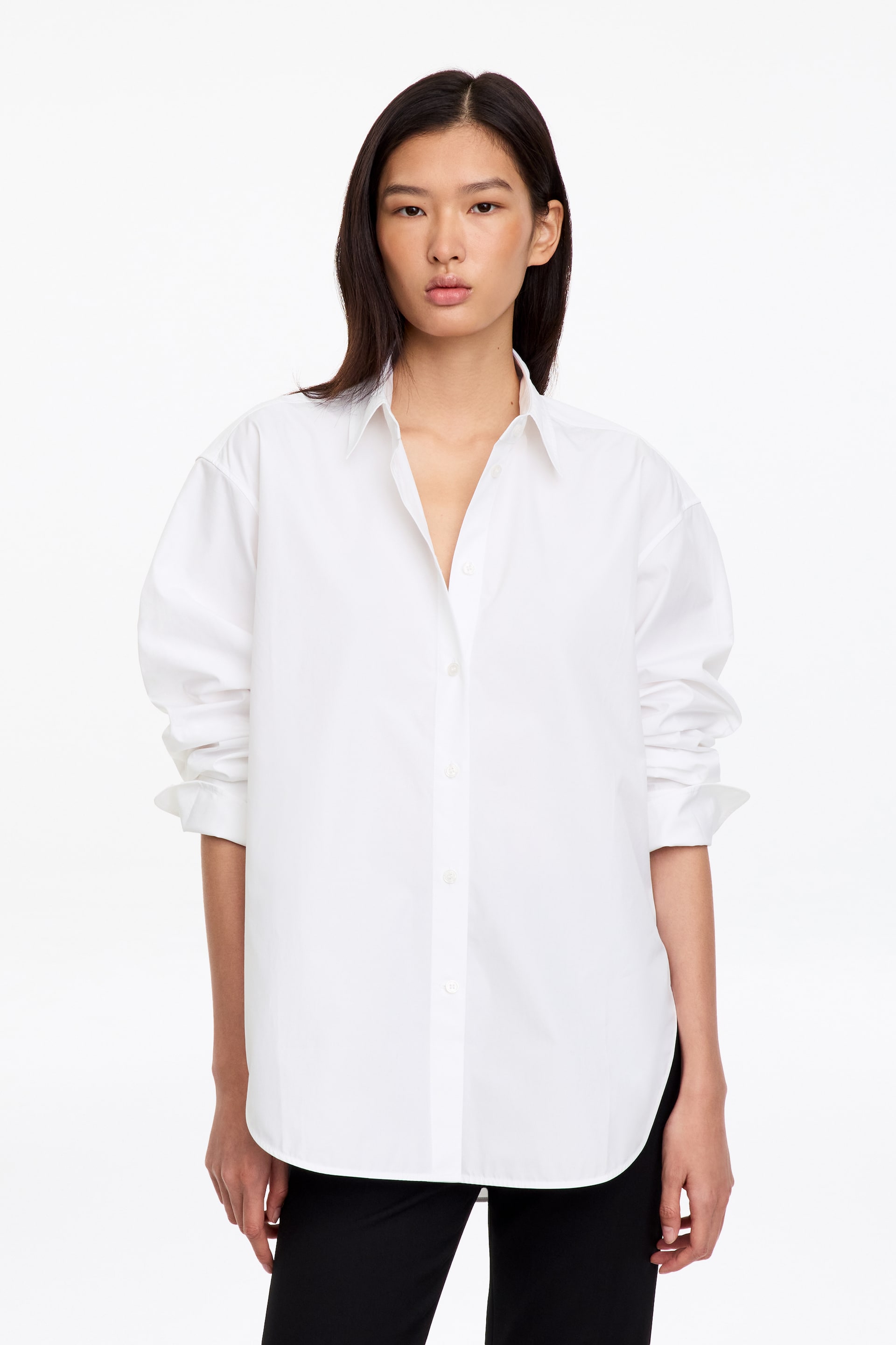 View larger image: A white, long-sleeved button-up shirt in crisp cotton poplin is worn by the model, featuring a relaxed silhouette, a pointed collar, and a curved hem. The shirt is styled with the top buttons undone and the sleeves slightly pushed up, paired with black trousers.