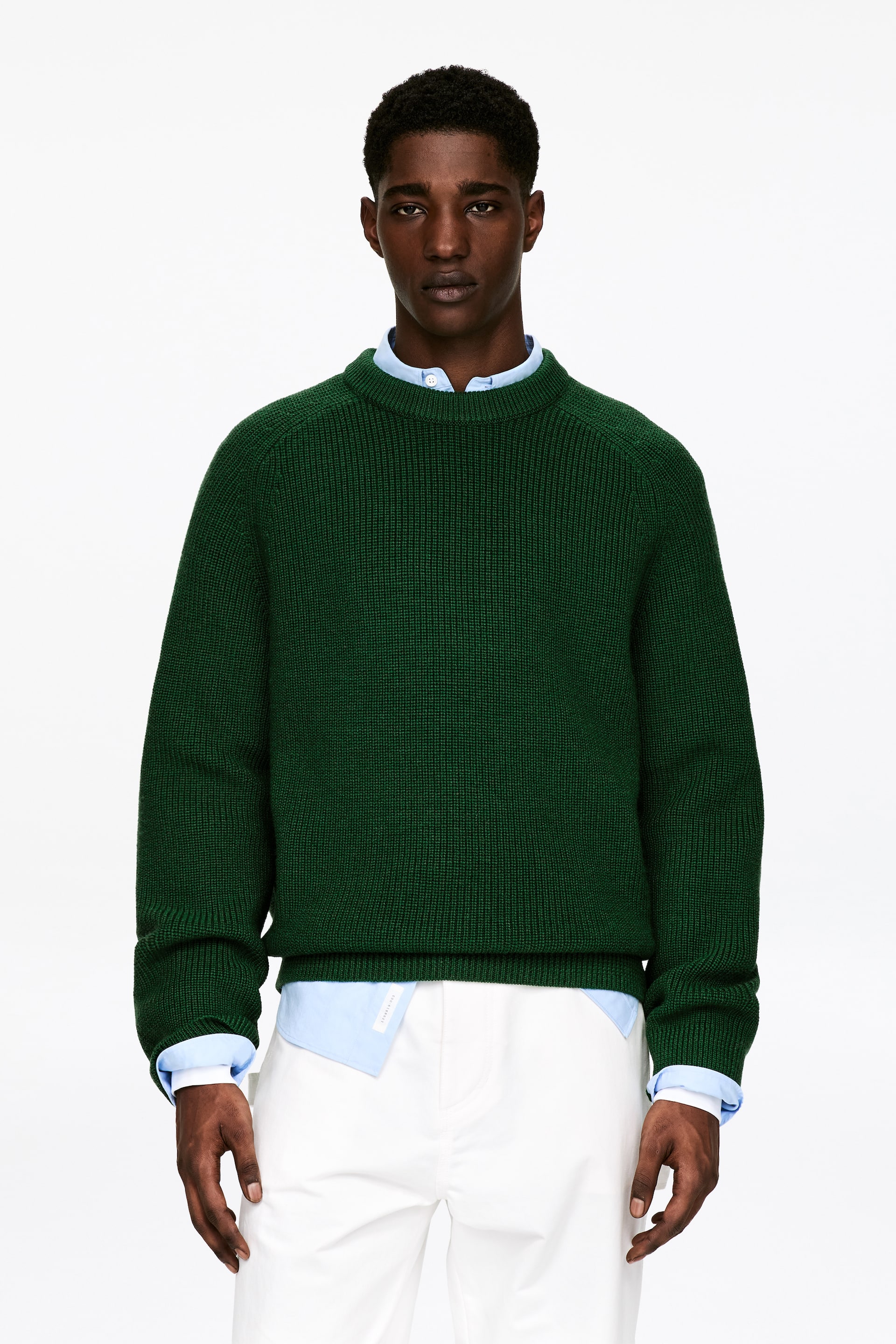 Größeres Bild ansehen: Ein dunkelgrüner Pullover wird präsentiert, der einen strukturierten Strickkörper, Raglanärmel, einen Rundhalsausschnitt sowie gerippte Bündchen und einen gerippten Saum aufweist. Er wird über einem hellblauen Hemd mit Kragen getragen, wobei der Kragen und die Manschetten des Hemdes sichtbar sind.
