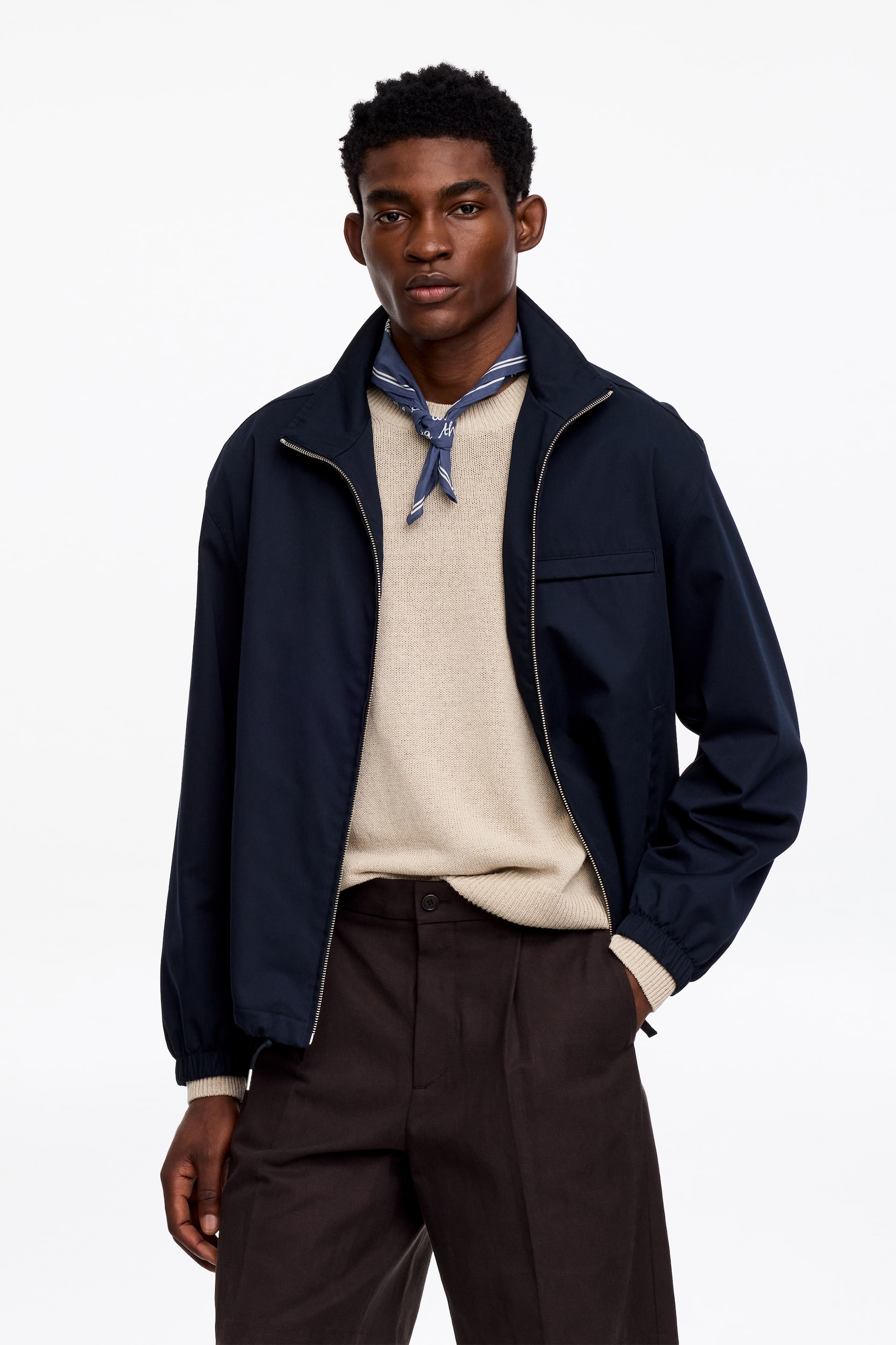 View larger image: A navy blue jacket featuring a stand-up collar, a full front zip, and ribbed cuffs. It is layered over a light beige jumper and a blue patterned bandana, with a flap pocket visible on the wearer's left chest.
