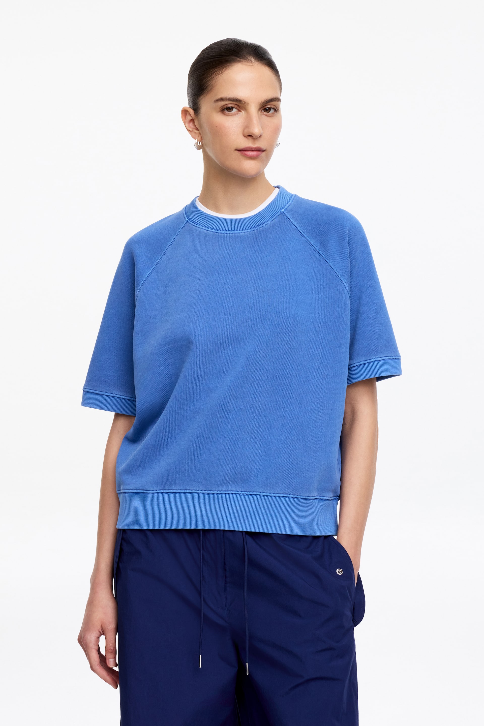 Se større billede: Den mellemblå kortærmede sweatshirt har en rund halsudskæring, raglanærmer og ribbede manchetter på de korte ærmer. En ribbet kant fuldender designet, og en hvid t-shirt er synlig under halsudskæringen.