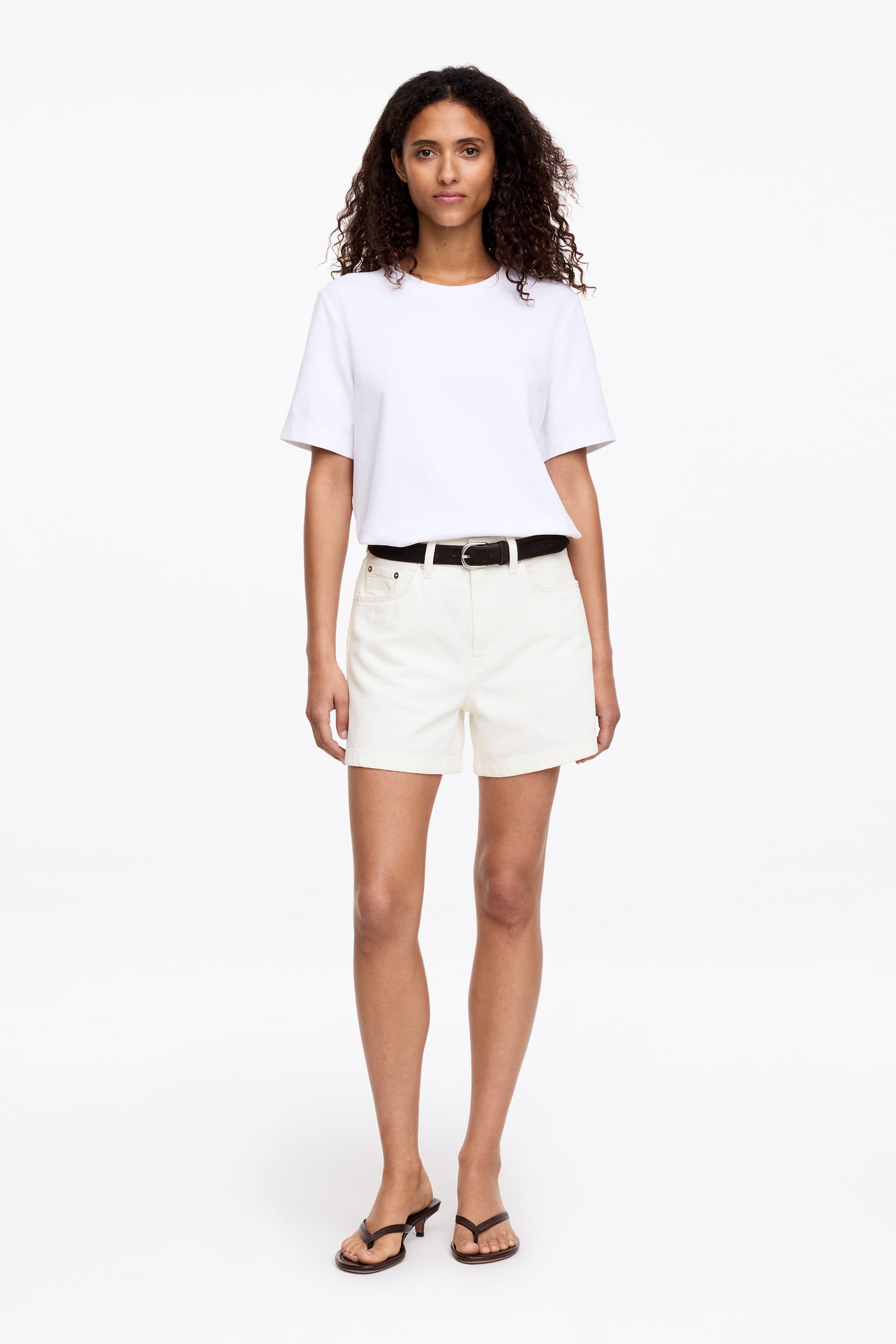 Agrandir l'image: Un t-shirt blanc est présenté de face, avec un col rond et des manches courtes à coupe décontractée. Son tissu lisse semble rentré dans un short de couleur claire, avec l'ourlet tombant à la taille.