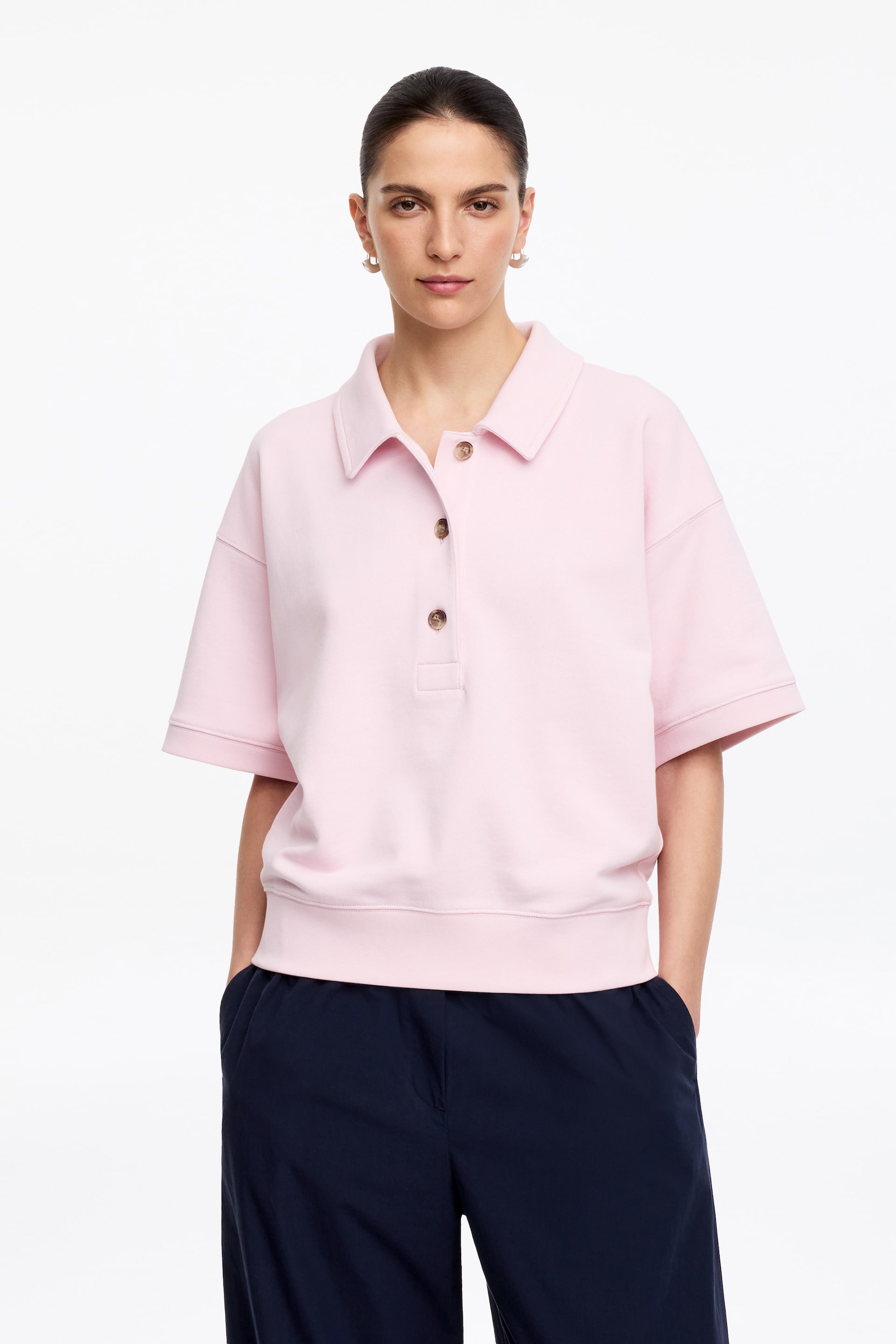 Visualizza immagine più grande: Una polo rosa a maniche corte viene presentata frontalmente, mostrando una vestibilità rilassata. Presenta un classico colletto polo, una chiusura a tre bottoni con bottoni marrone chiaro, maniche corte che raggiungono il gomito e un orlo con fascia.