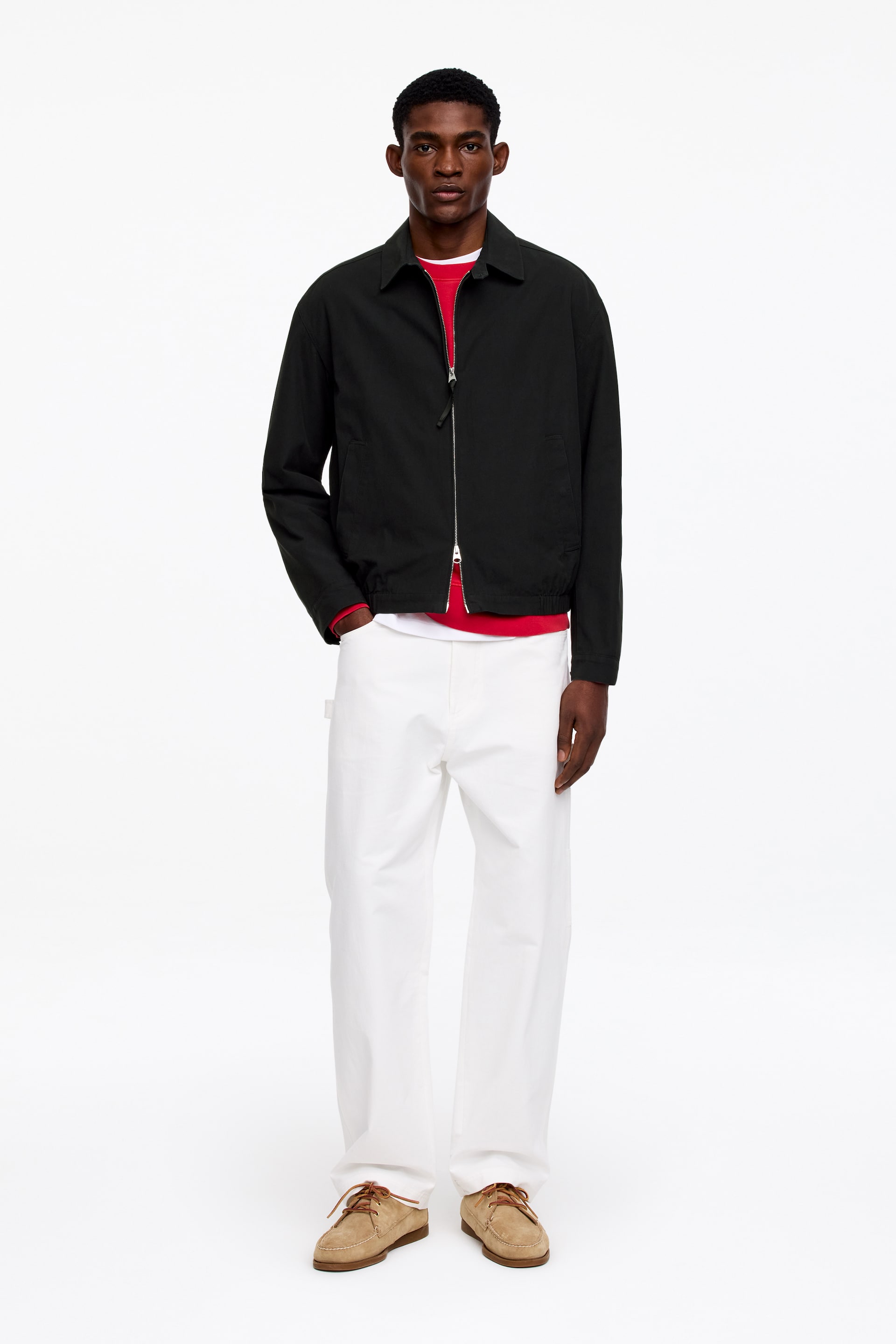 Zobacz większy obraz: Czarna kurtka prezentowana jest z widoku frontalnego, z klasycznym szpiczastym kołnierzem i srebrnym zamkiem błyskawicznym na całej długości. Jest noszona rozpięta, odsłaniając czerwoną koszulkę z długim rękawem pod spodem i eksponując dwie ukośne kieszenie z wypustkami z przodu.