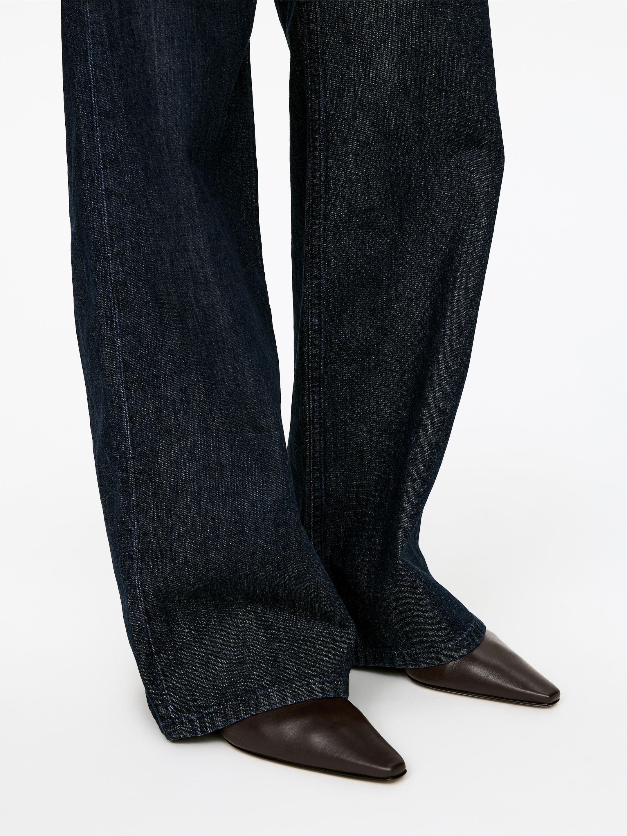 Grotere afbeelding bekijken: Donker gewassen denim jeans tonen een rechte, ontspannen pasvorm met subtiele vervaging langs de naden, vallend over donkerbruine schoenen met puntige neus.