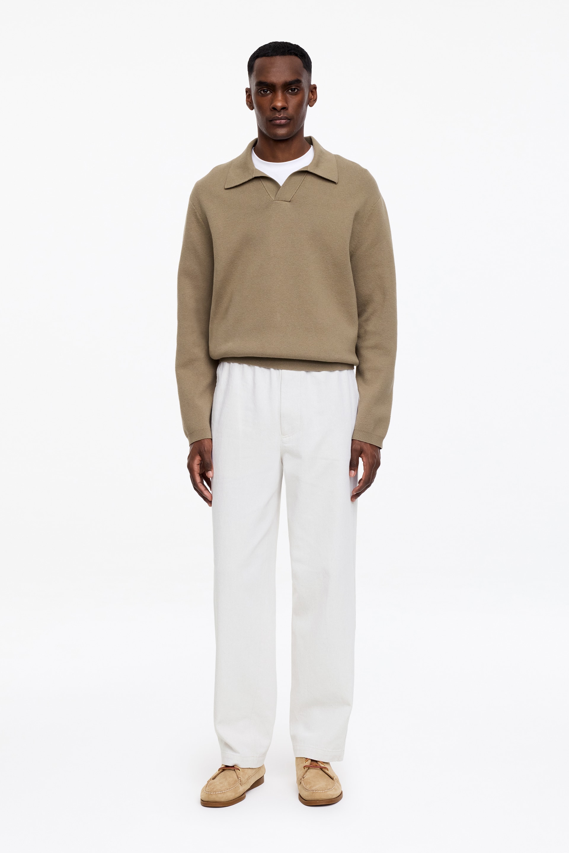 Vis større bilde: En tanfarget strikket polosweater vises fra forsiden, med en spredt krage, en V-halsåpning, lange ermer med ribbestrikkede mansjetter og en ribbestrikket kant, med en hvit t-skjorte med rund hals synlig under.