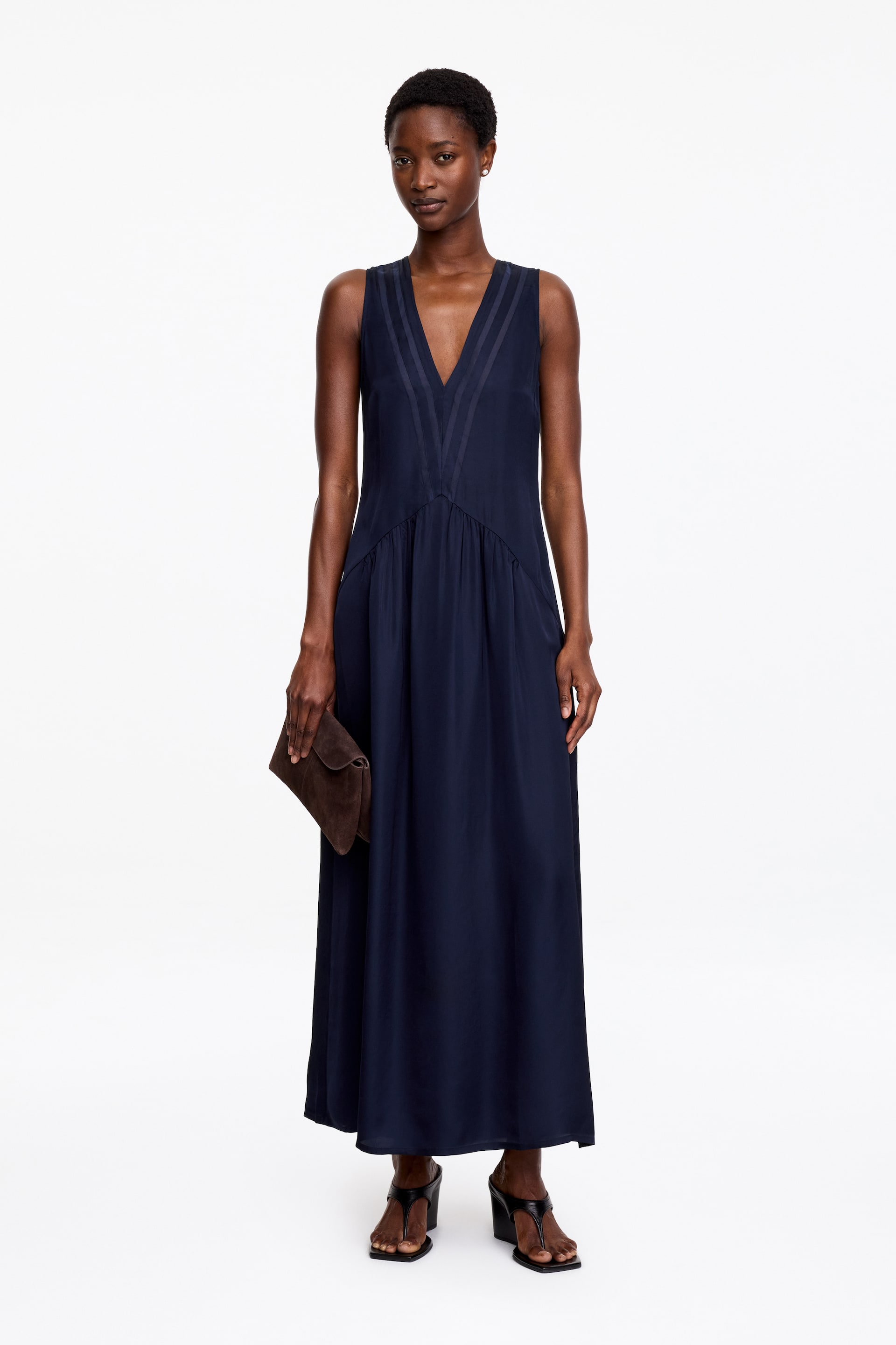 Agrandir l'image: La mannequin porte une robe longue sans manches bleu marine, avec des insertions en organza de soie au niveau du buste et des détails froncés sur la jupe, créant un drapé doux et fluide. Le modèle tient une pochette marron et porte des sandales à talons hauts noires.