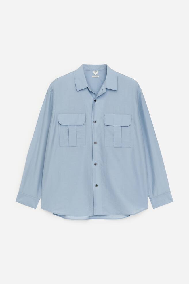 A light blue long-sleeved shirt is showcasing its front, featuring a spread collar, a full button-front fastening with dark buttons, and two large chest pockets with flaps and pleats. - 1