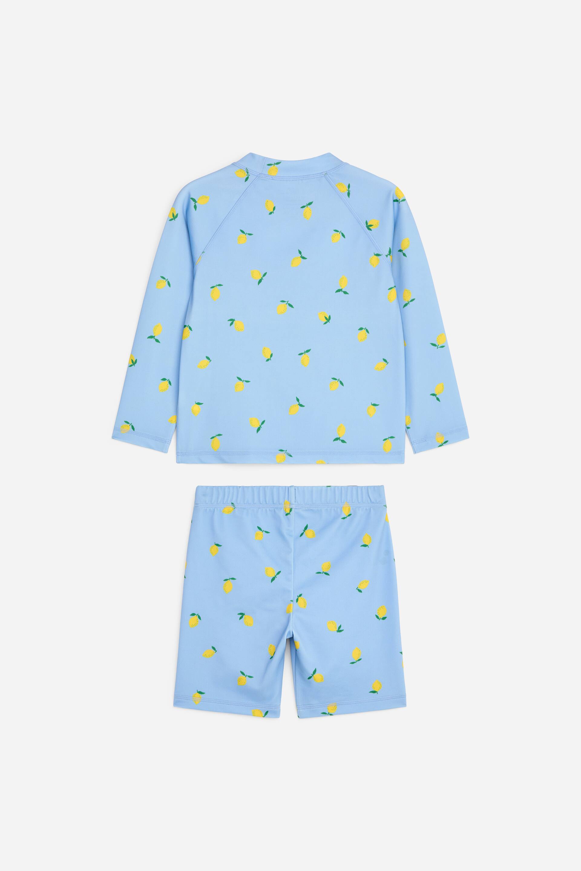 Grotere afbeelding bekijken: Een lichtblauwe zwemsset wordt van de achterkant getoond, bestaande uit een longsleeve t-shirt met een opstaande kraag en raglanmouwen, en knielange broekjes, beide versierd met een herhalend patroon van gele citroenen; de broekjes hebben ook een elastische tailleband.