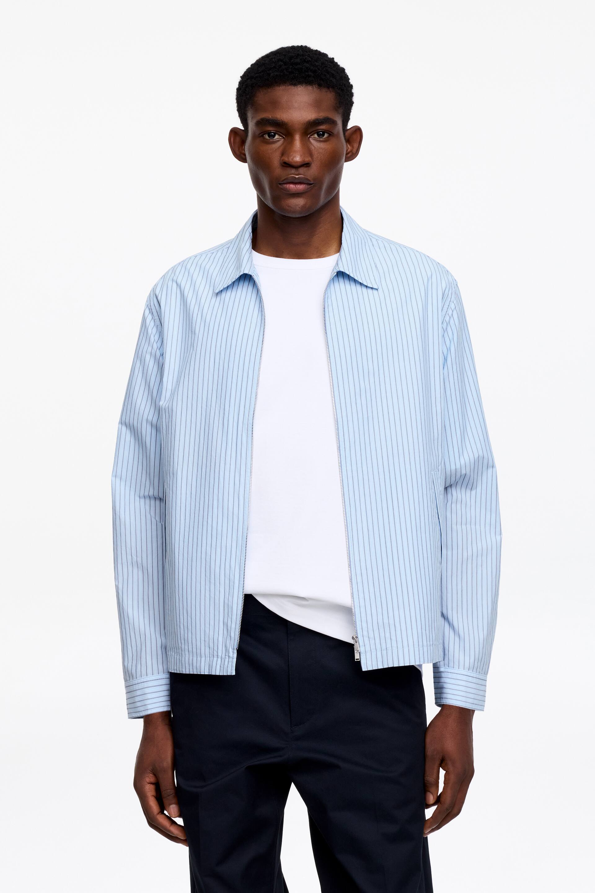 Zobrazit větší obrázek: Světle modrá pruhovaná bunda s rozšířeným límcem, dlouhými rukávy s manžetami na knoflíky a stříbrným zipem. Bunda je částečně rozepnutá přes bílé tričko, což zdůrazňuje její uvolněný střih a rovný lem.