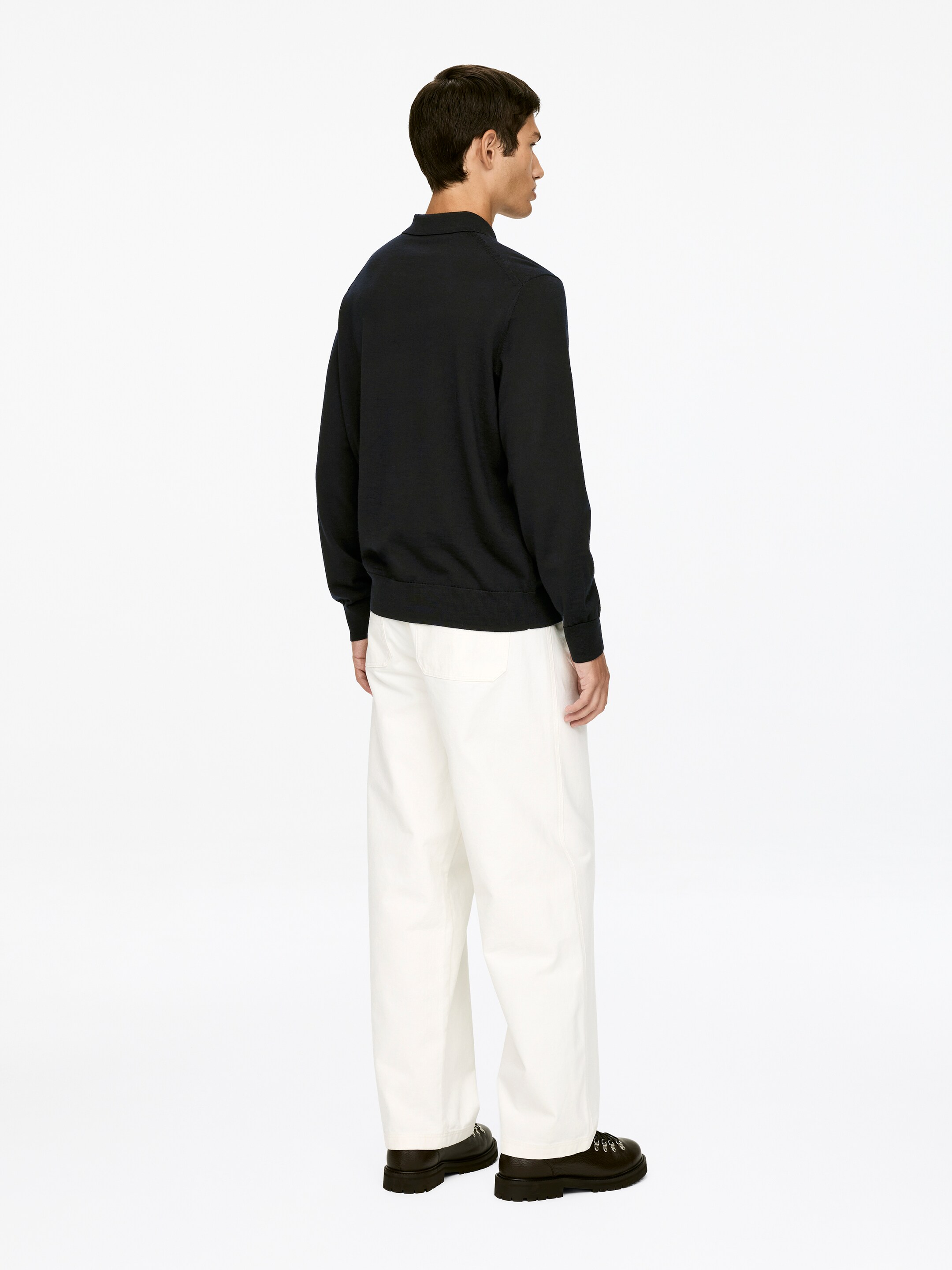 Visualizza immagine più grande: Una polo a maniche lunghe di un blu scuro con colletto e polsini a coste viene indossata con pantaloni a gamba larga color avorio. Il taglio rilassato della maglietta e la larghezza dei pantaloni sono visibili dall'angolazione posteriore, con i pantaloni che cadono su scarpe scure e robuste con lacci.