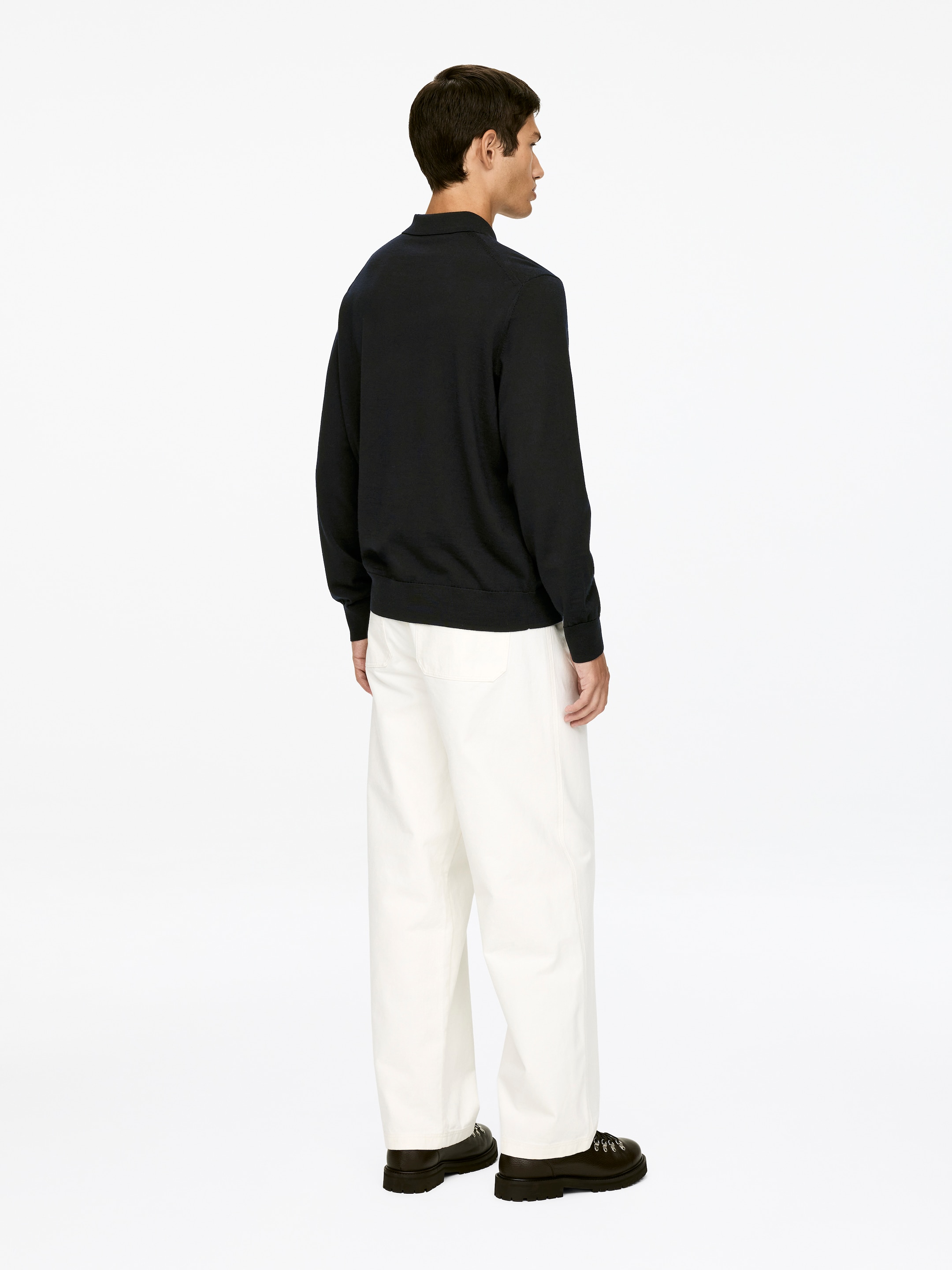 Grösseres Bild ansehen: Ein dunkelblaues Langarm-Poloshirt mit Kragen und gerippten Bündchen wird mit off-white, weit geschnittenen Hosen getragen. Der lockere Sitz des Shirts und die Weite der Hosen sind aus dem Blickwinkel von hinten sichtbar, wobei die Hosen über klobige, dunkle Schnürschuhe fallen.