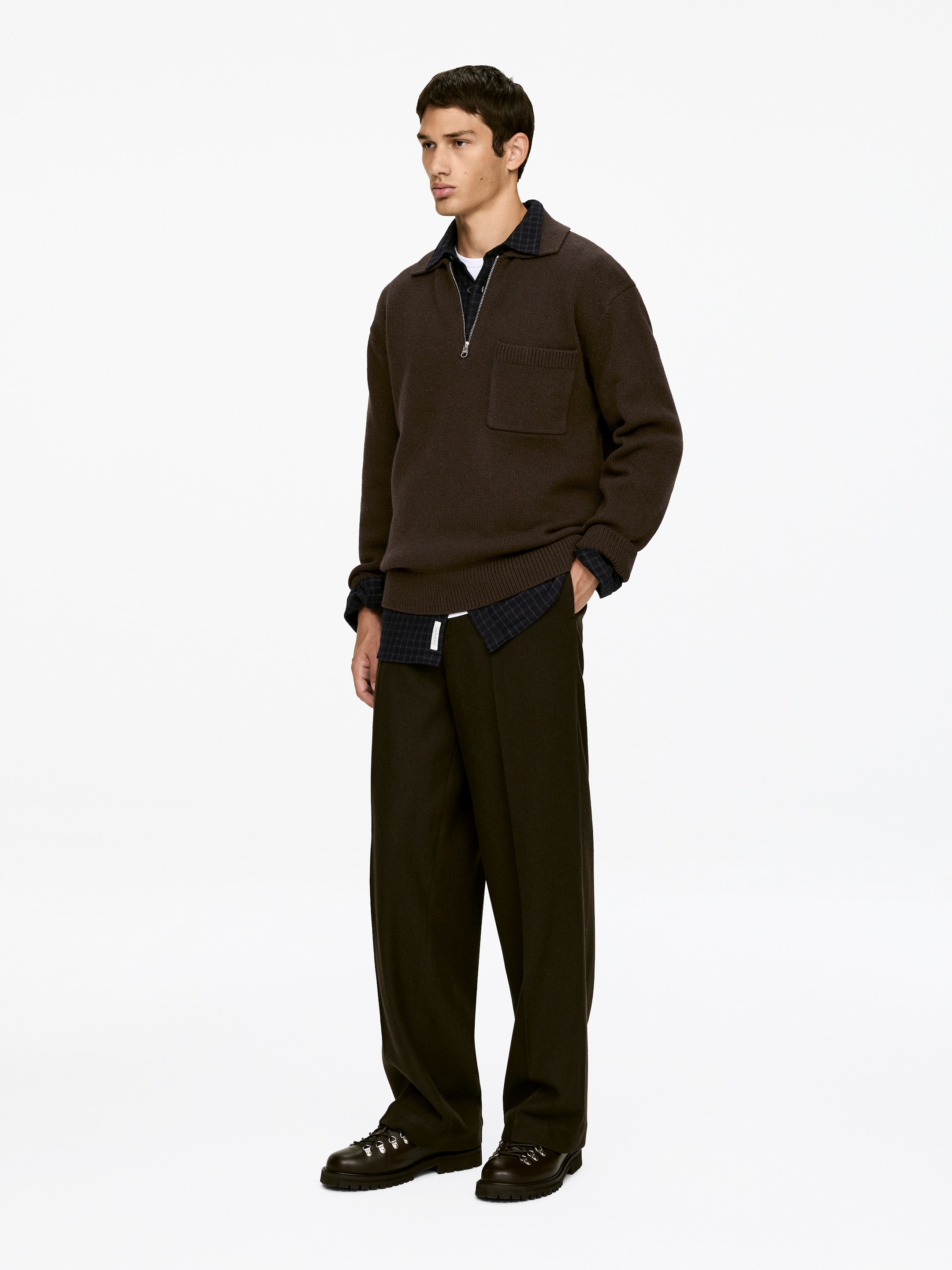 Größeres Bild anzeigen: Ein Outfit wird in einer Dreiviertelansicht präsentiert, mit einem braunen Strick-Halbzipp-Pullover mit einem ausgestellten Kragen, einem silbernen Reißverschluss und einer Brusttasche auf der linken Seite, der über einem dunklen Karohemd und einem weißen T-Shirt liegt. Das Ensemble wird durch weit geschnittene braune Hosen vervollständigt, die eine weiße Kordel zeigen, und dunkle Schnürstiefel mit D-Ring-Ösen und einer Profilsohle.