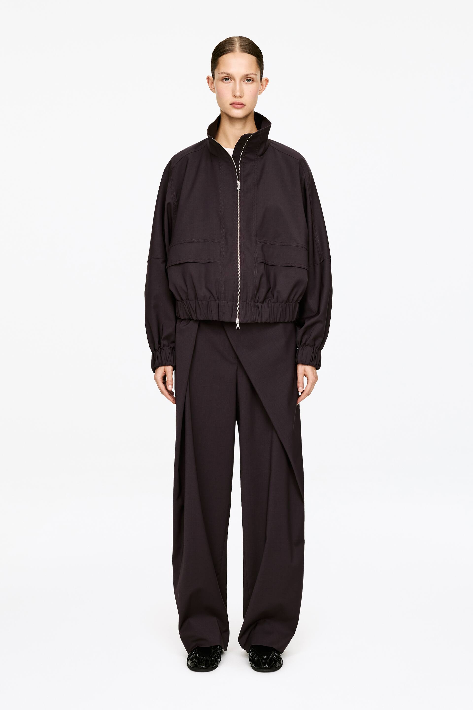 Ver imagen más grande: Una chaqueta blusón de color carbón oscuro y pantalones de pierna ancha a juego se presentan de frente. La chaqueta cuenta con un cuello alto, una cremallera plateada en la parte delantera, dos bolsillos con solapa en el pecho y un dobladillo y puños elásticos, llevada sobre una camiseta blanca. Los pantalones están diseñados con un panel frontal superpuesto.