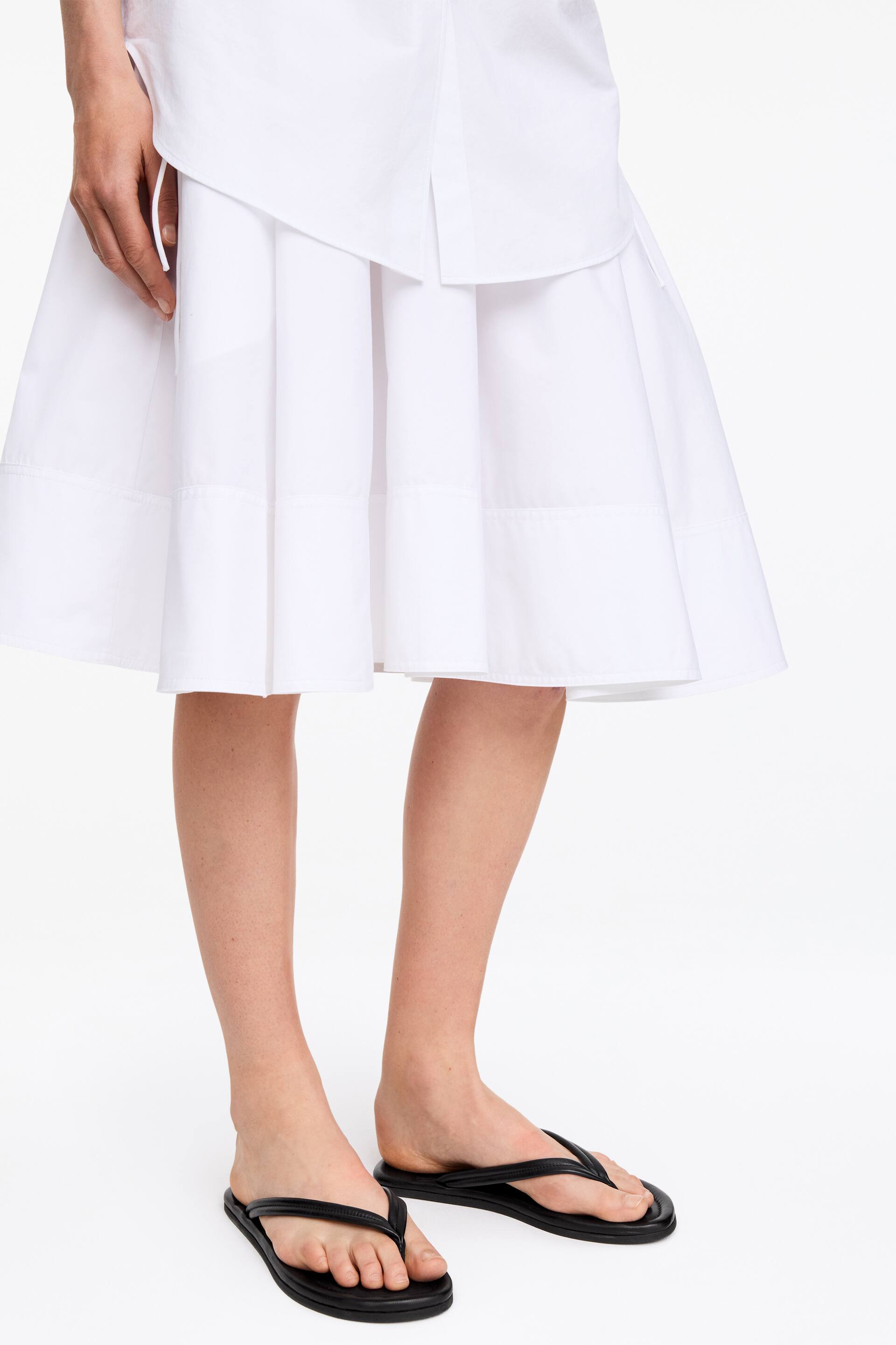 Visa större bild: En vit rynkad kjol presenterar en voluminös silhuett och har en bred horisontell panel ovanför fållen. En vit skjorta lager över kjolens övre del, vilket avslöjar dess flödande drapering.