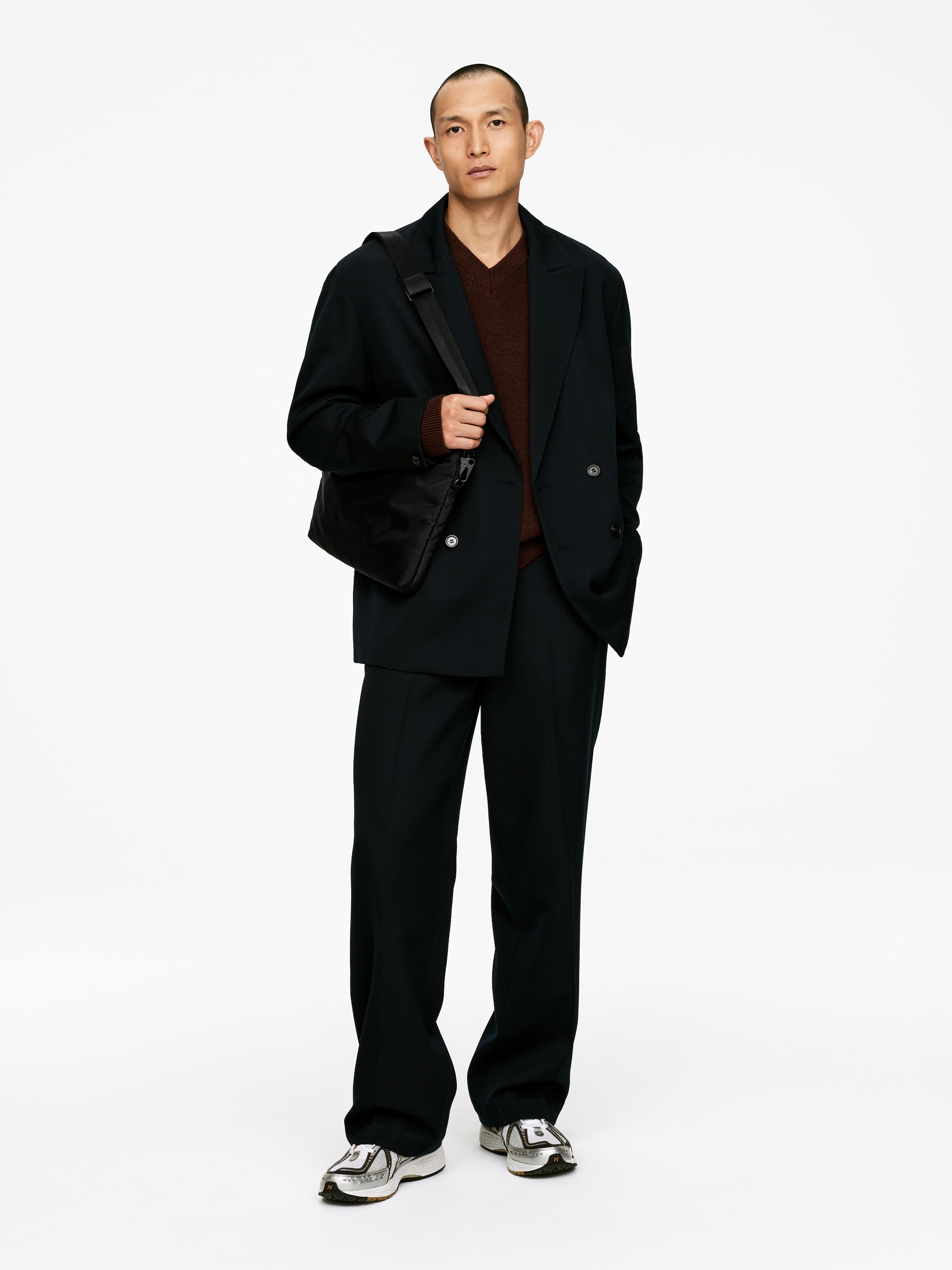Agrandir l'image: Un costume surdimensionné bleu marine foncé est porté, face à l'avant, avec un blazer croisé à larges revers en pointe et un pantalon assorti à jambes larges qui s'accumulent aux chevilles. Un pull marron à col en V est superposé sous le blazer, tandis qu'un sac à bandoulière noir est porté sur l'épaule et des baskets argentées sont portées.