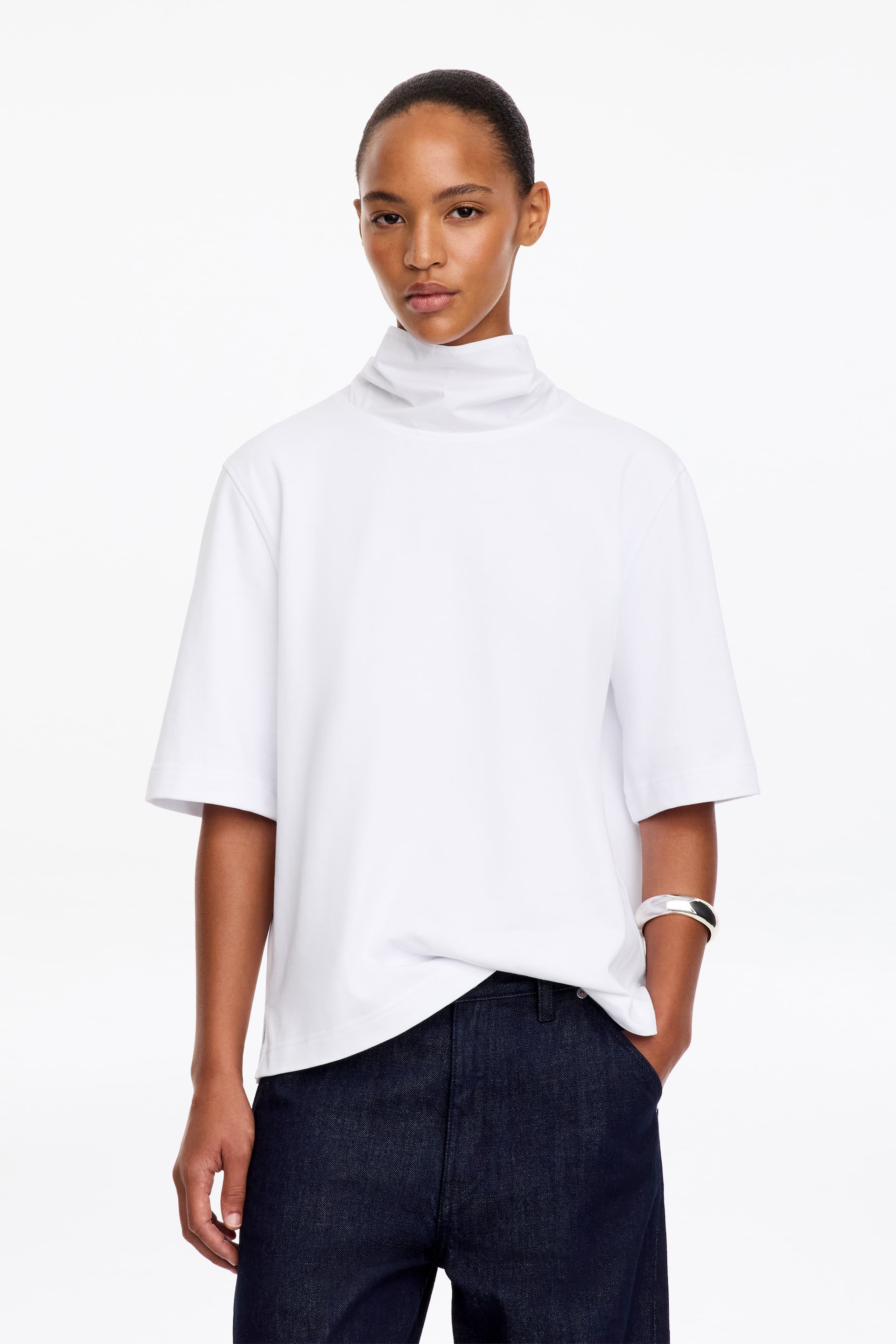 Visualizza l'immagine ingrandita: Un top bianco dalla vestibilità regolare, con collo alto e dettaglio foulard drappeggiato e maniche a tre quarti, indossato dalla modella. Il top è abbinato a pantaloni in denim scuro e a un bracciale rigido argentato.