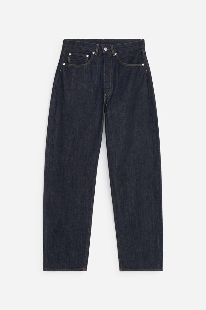 Een donkergewassen indigoblauwe denimjeans staat naar voren gericht en toont een relaxte pasvorm met barrel-vormige pijpen en een mid-rise taille. De jeans heeft een klassieke five-pocket-styling met oranje contrasterende stiksels en zilverkleurige rivets. - 1