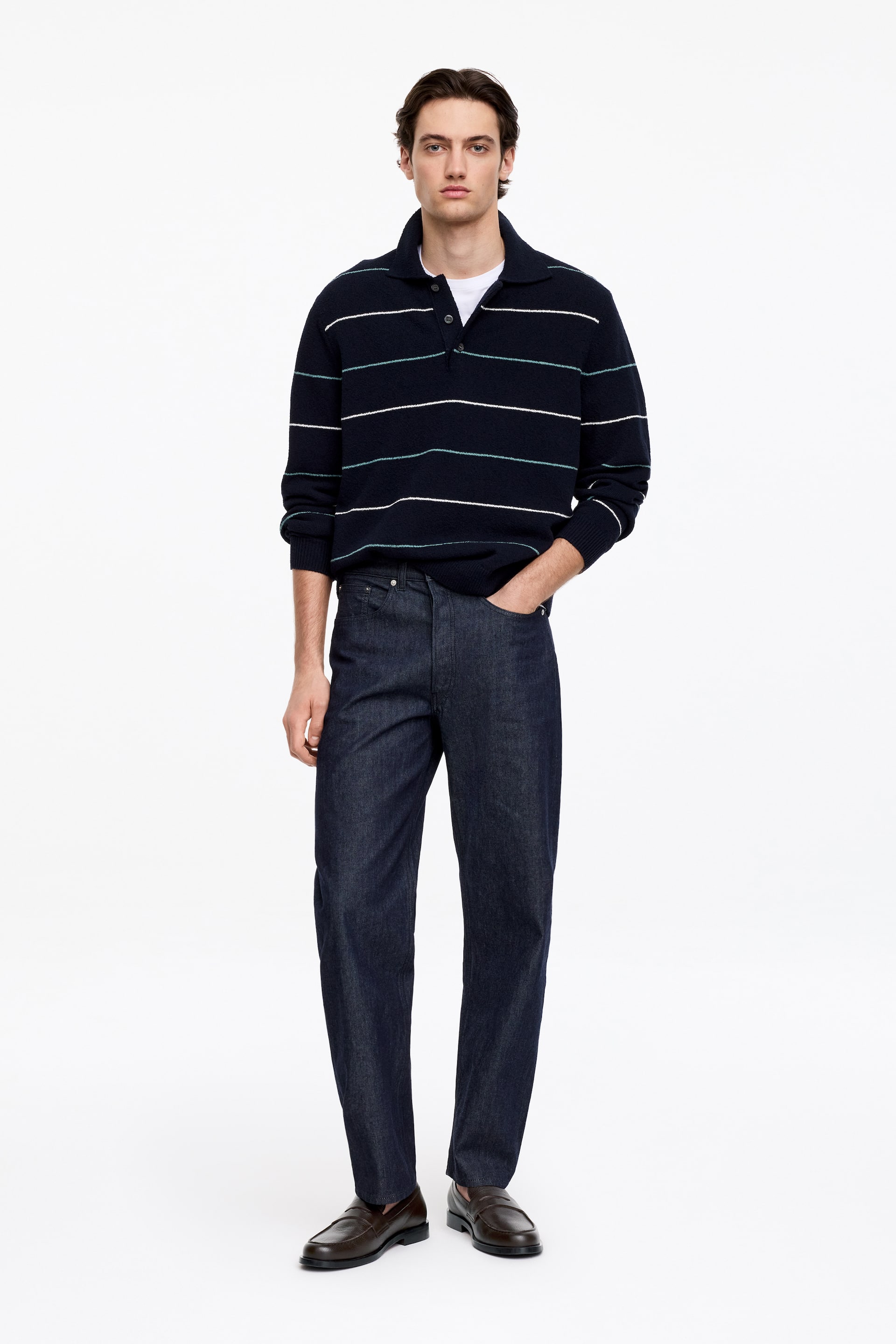 Größeres Bild ansehen: Ein marineblauer gestreifter Polopullover, dunkel gewaschene Jeans und braune Loafer werden an einer Person präsentiert, die nach vorne blickt. Der Pullover zeichnet sich durch weiße und mintgrüne horizontale Streifen, einen Kentkragen und eine Dreiknopfleiste aus, getragen über einem weißen T-Shirt. Die dunkel gewaschenen Jeans zeigen eine gerade Passform und die braunen Loafer sind im klassischen Slipper-Stil gehalten.