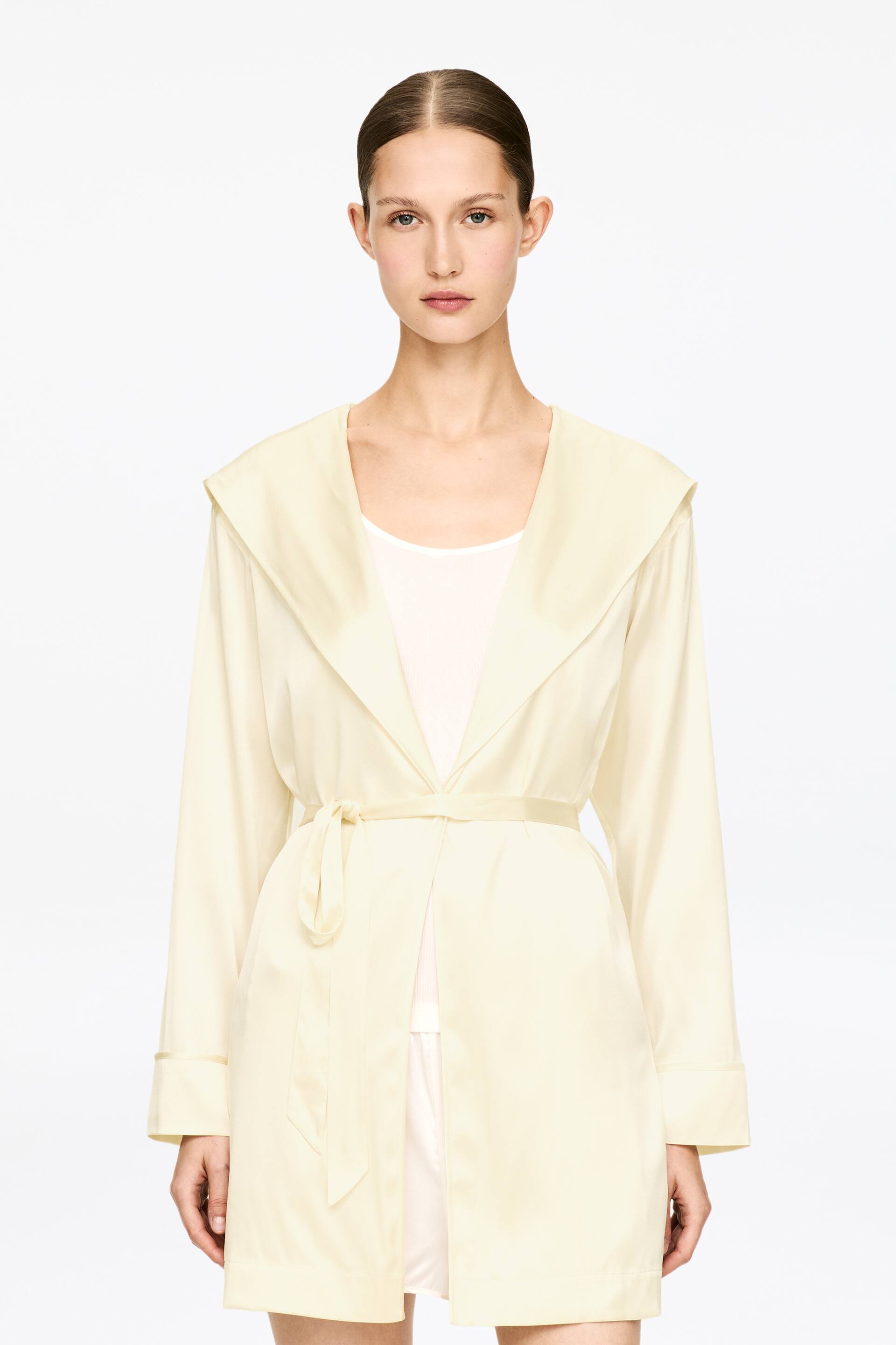 Visa större bild: En krämfärgad kort morgonrock med en subtil glans bärs av modellen. Den har en sjalkrage som övergår i en huva, och dess knytbälte är knutet i midjan över en ljusfärgad topp och shorts.