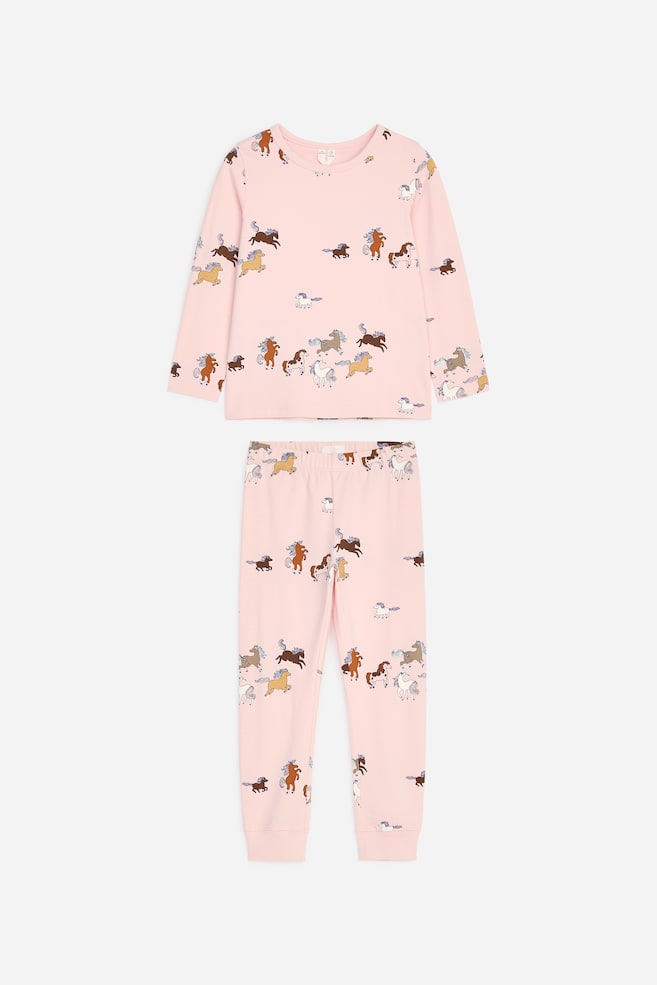 Un pigiama per bambini in due pezzi, di colore rosa chiaro, è mostrato frontalmente e comprende una maglia a maniche lunghe con girocollo e un pantalone lungo coordinato con polsini a coste alla caviglia. Entrambi i capi presentano una stampa all-over di cavalli al galoppo multicolori. - 1