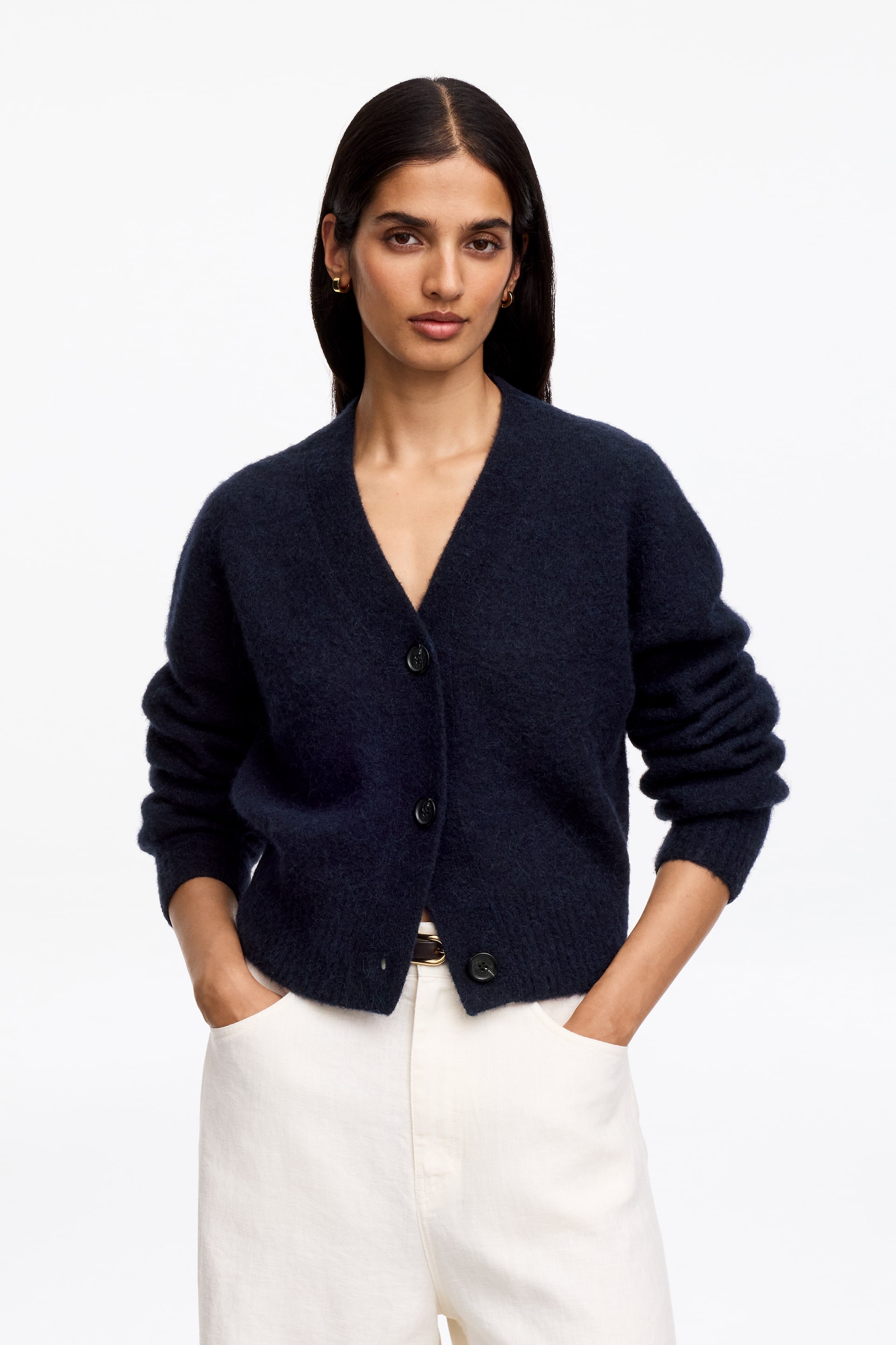 Grotere afbeelding bekijken: Een alpaca-wolblend vest in marineblauw wordt gedragen door het model. Het pluizige, v-hals vest, met drie donkere knopen, is gedeeltelijk dichtgeknoopt en gestyled over lichtgekleurde broeken, terwijl de handen van het model in de broekzakken rusten.