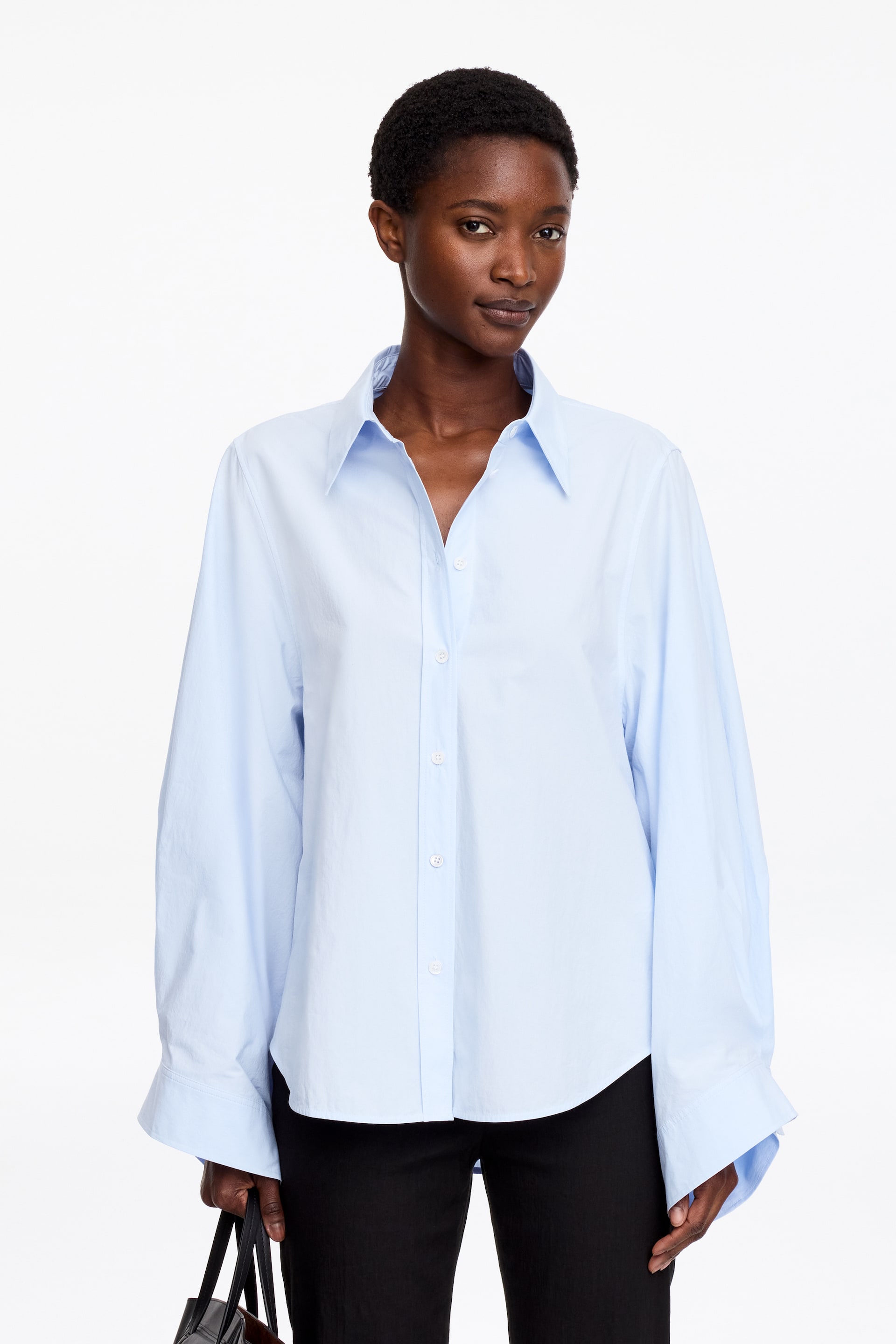 Agrandir l'image: Une chemise en coton bleu clair avec un col classique et une fermeture boutonnée est portée par le mannequin. Elle se distingue par ses manches subtilement volumineuses avec des poignets larges, et le mannequin tient un sac noir.