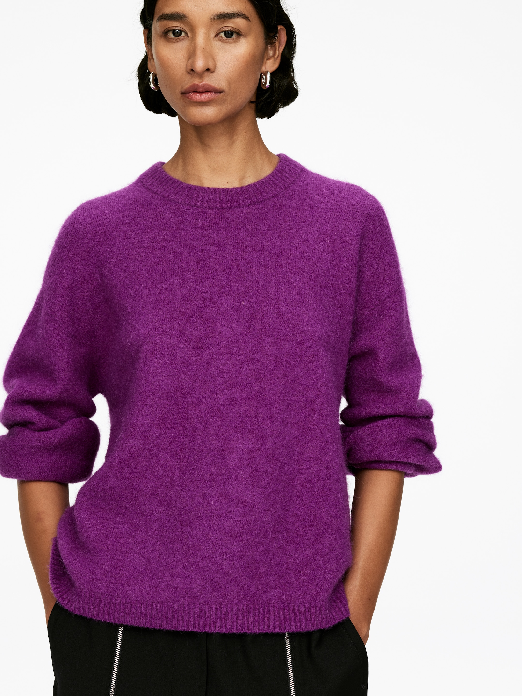 Agrandir l'image: Un pull à col rond violet vif est porté, avec une texture douce et duveteuse et une coupe décontractée. Ses manches longues sont retroussées aux poignets, et son ourlet côtelé est légèrement tiré par des mains dans les poches.
