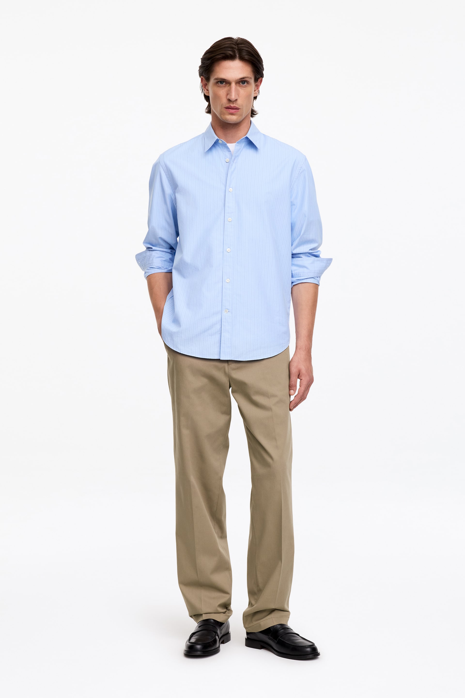 Ver imagen más grande: La modelo lleva una camisa abotonada a rayas finas de color azul claro, por fuera del pantalón. Las mangas largas de la camisa están remangadas y se combina con pantalones caqui.