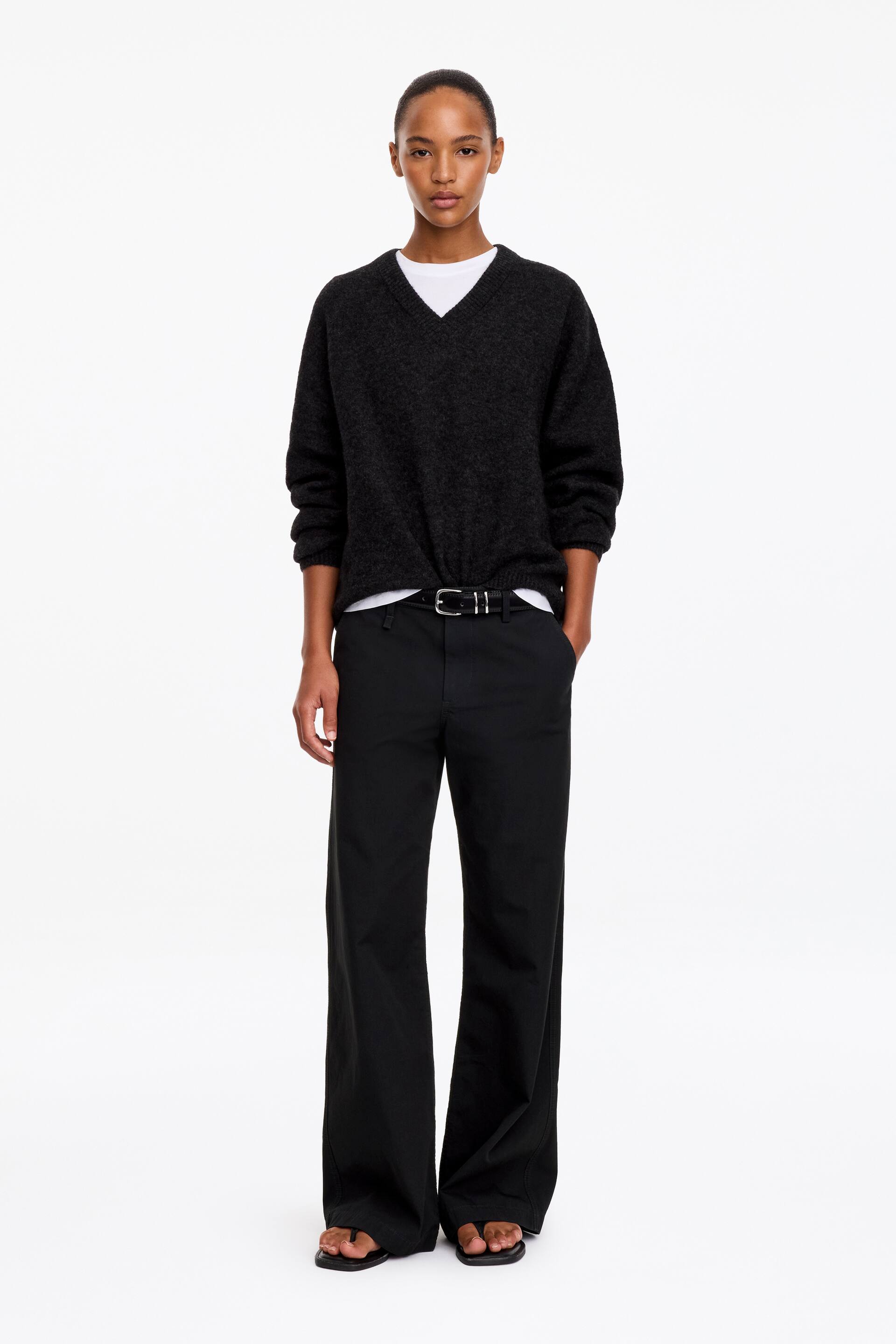 Visualizza l'immagine ingrandita: Il modello indossa pantaloni chino neri a gamba larga, con una mano appoggiata nella tasca laterale. I pantaloni sono abbinati a una cintura in pelle nera, a un maglione scollato a V grigio scuro sovrapposto a una T-shirt bianca e a sandali neri.
